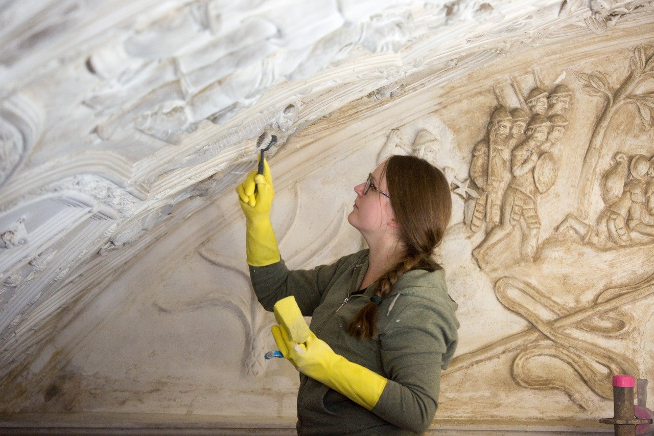 A conservator wearing yellow rubber gloves using a brush to clean the plaster on the ceiling in the Long Gallery at Lanhdyrock