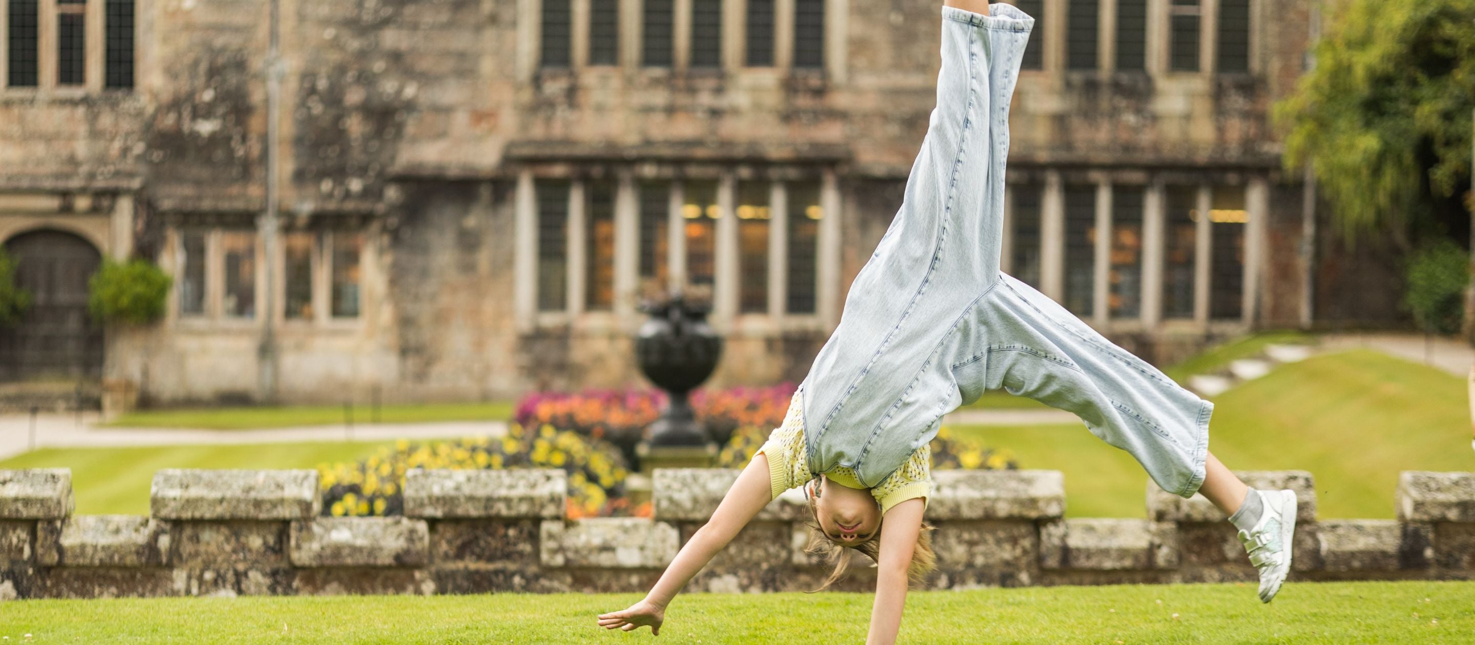 A child cartwheels across the lawn in front of Lanhydrock House on a bright summer day.