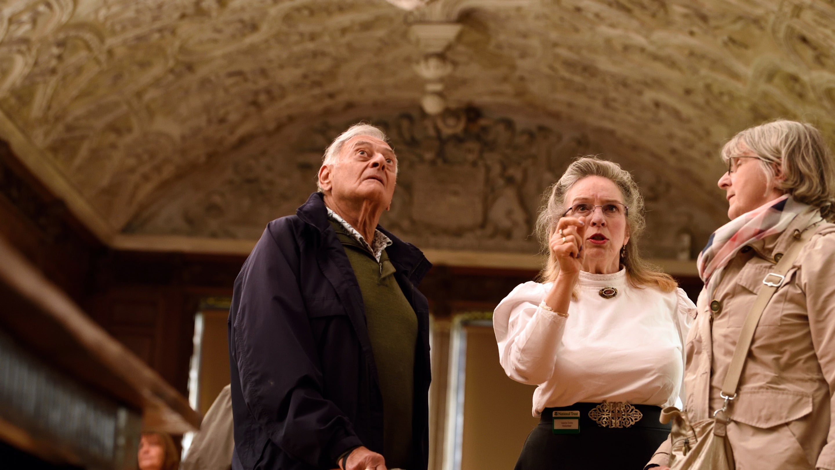 Two visitors and a room guide stood inside looking up at a plaster curved ceiling