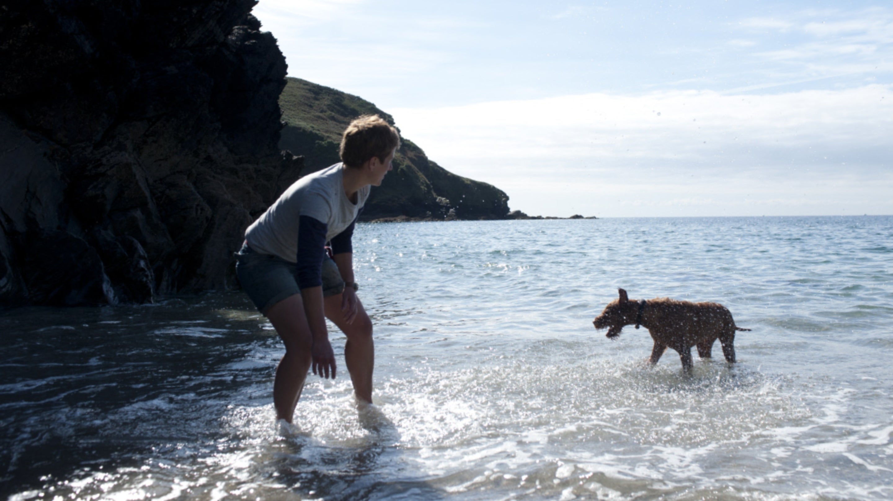 A person in the sunny shallows of the sea with their dog, with dark cliffs behind