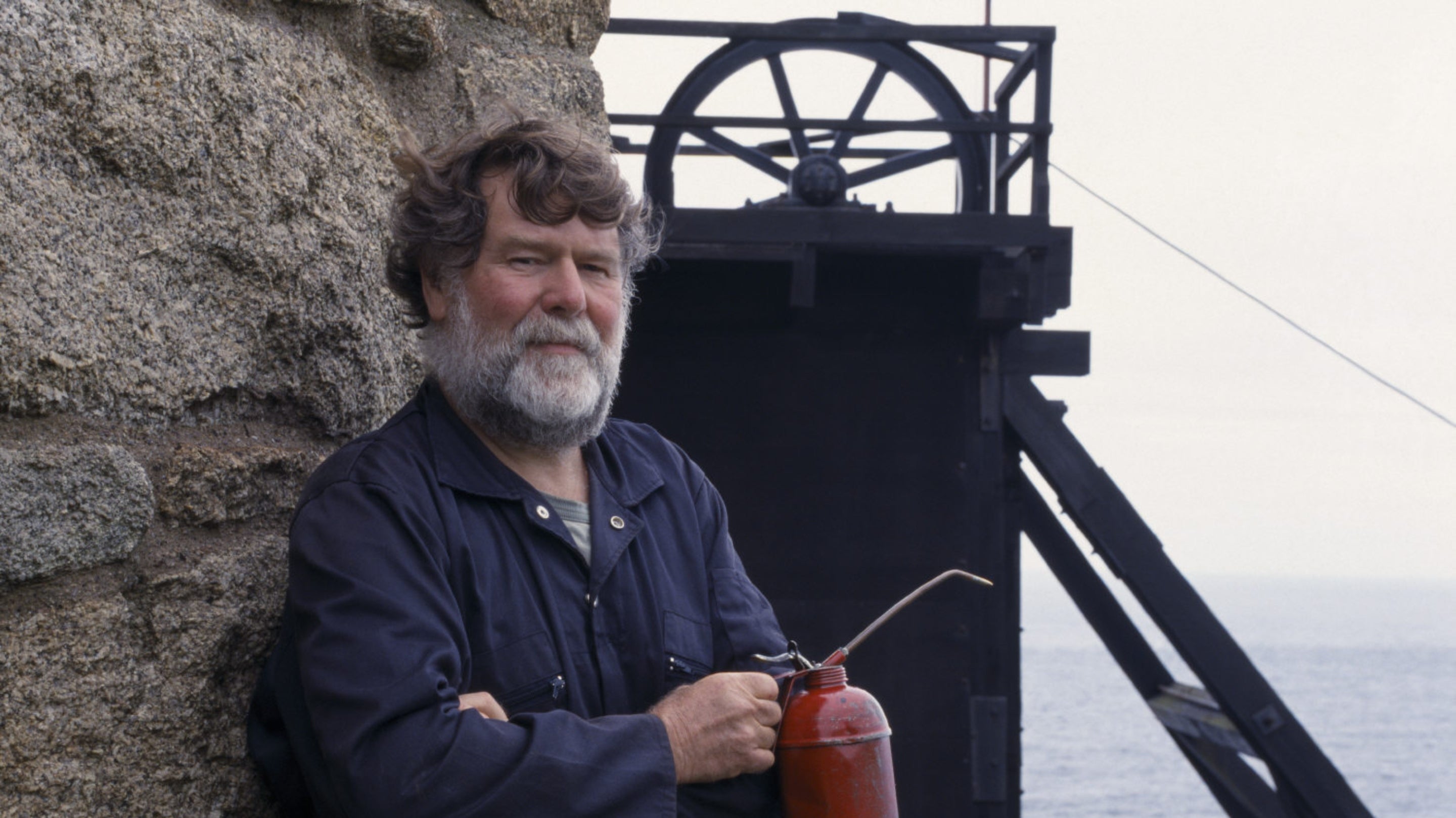 Volunteer Chris Quick is wearing a navy blue boiler suit and holding a red oil can, standing in front of a black wooden mine structure at Levant