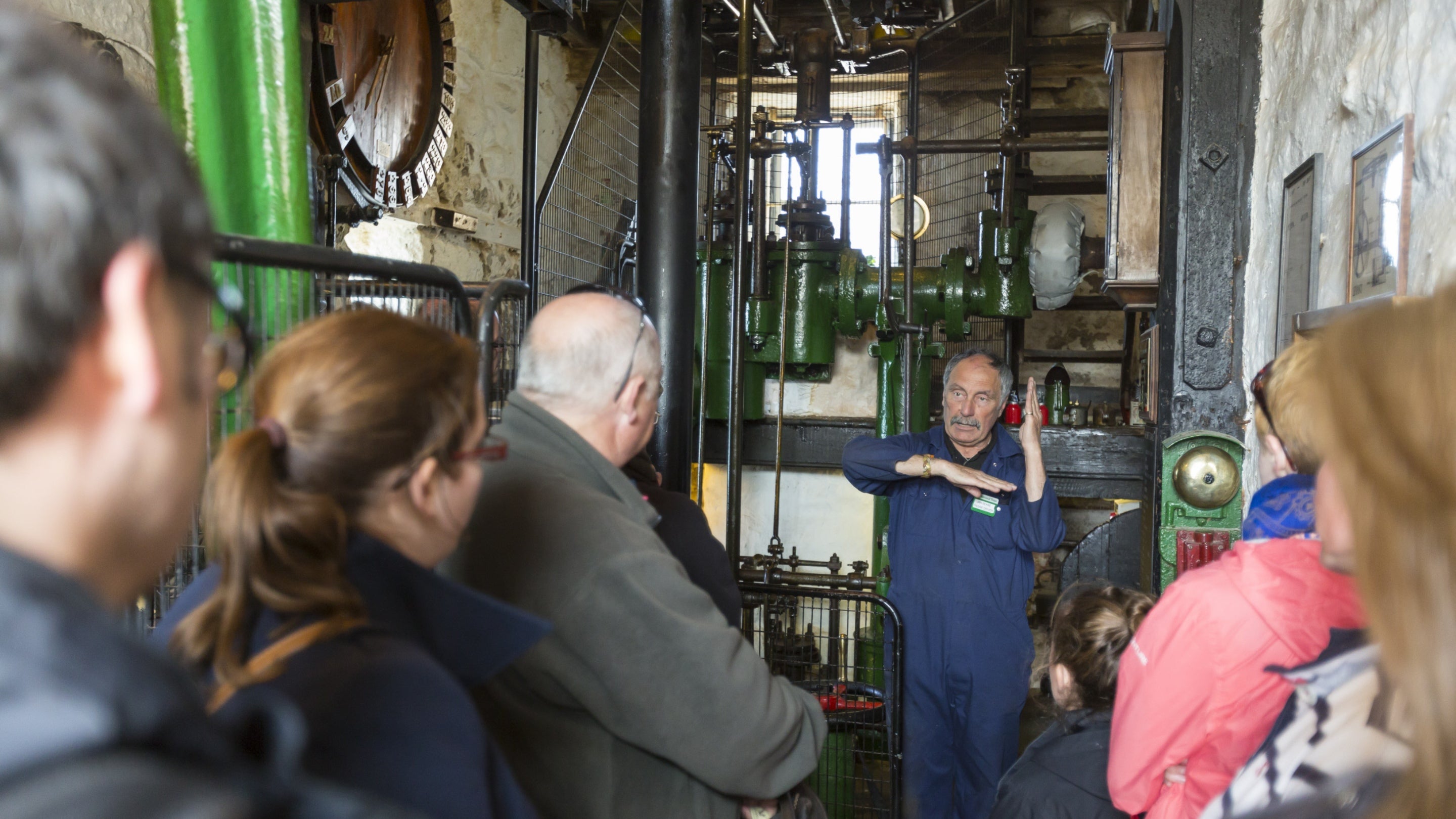 Visitors meet the volunteer who drives the beam engine at Levant Mine and Beam Engine, Cornwall. The volunteer stands in front of the machinery using his arms to show how it works.