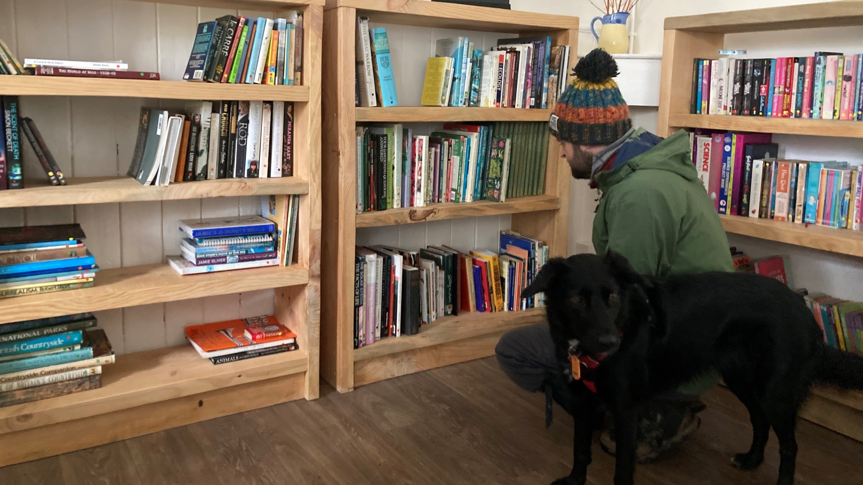 A visitor in a green coat and woolly hat browses the books, next to a black dog.