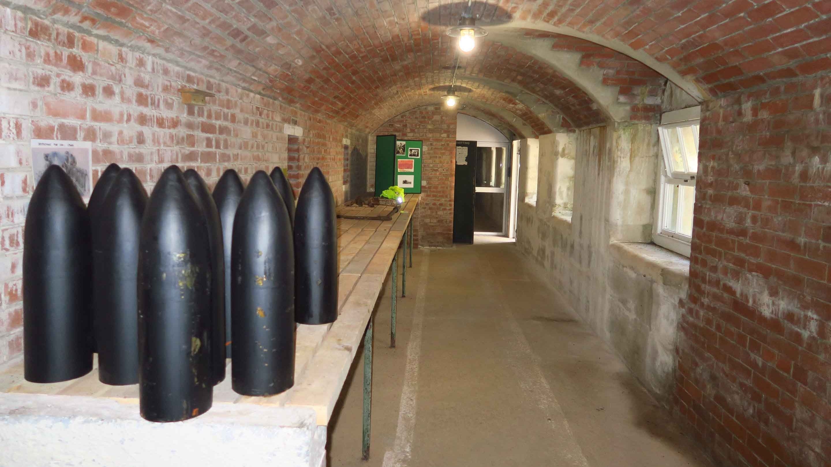 A brick corridor with a barrel ceiling and single bulb lights running down the middle of it. There are small windows in the external wall on the right. In the foreground gun ammunition can be seen.