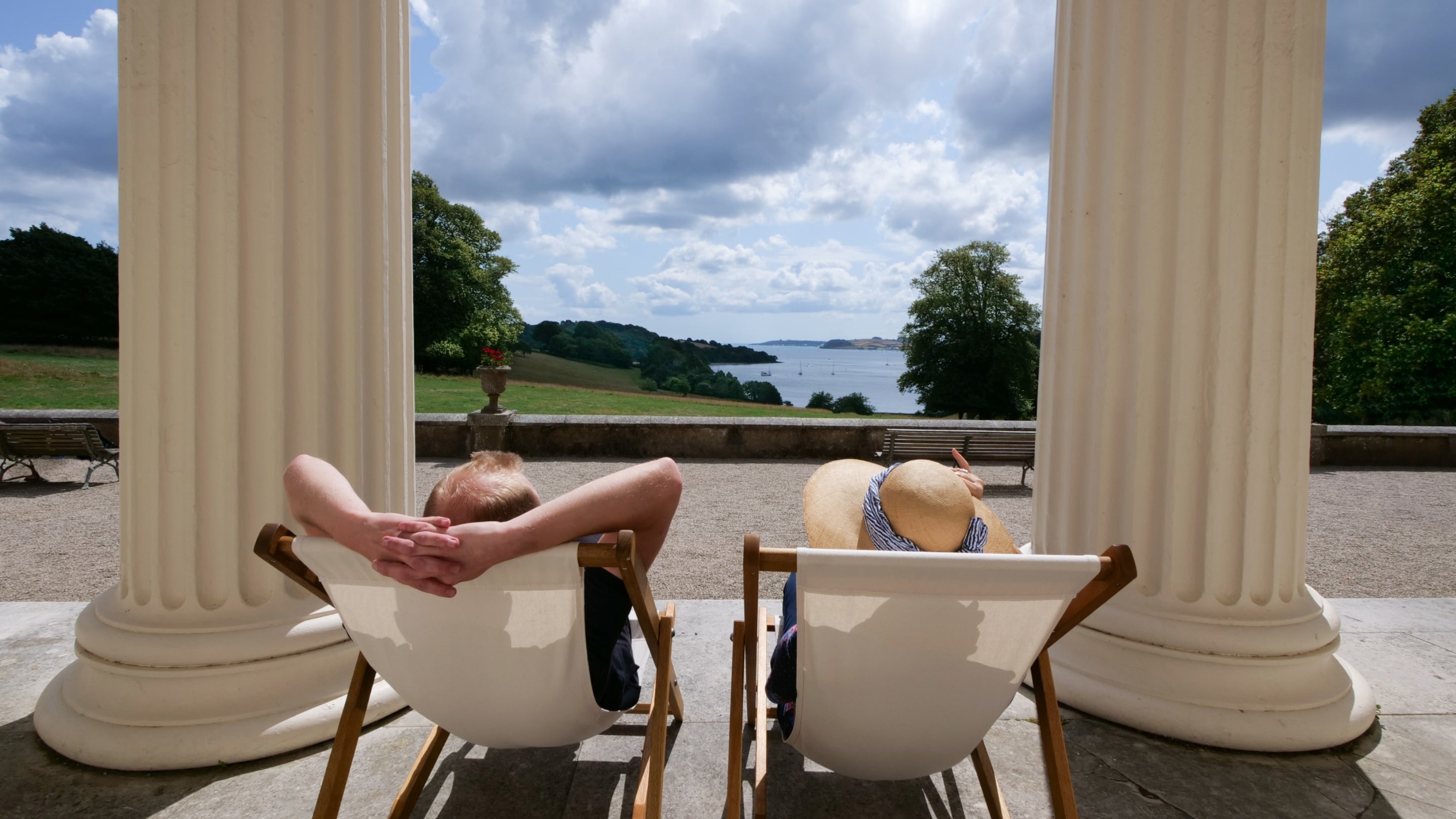 Two people sit in deckchairs on a colonnade terrace, looking out over water