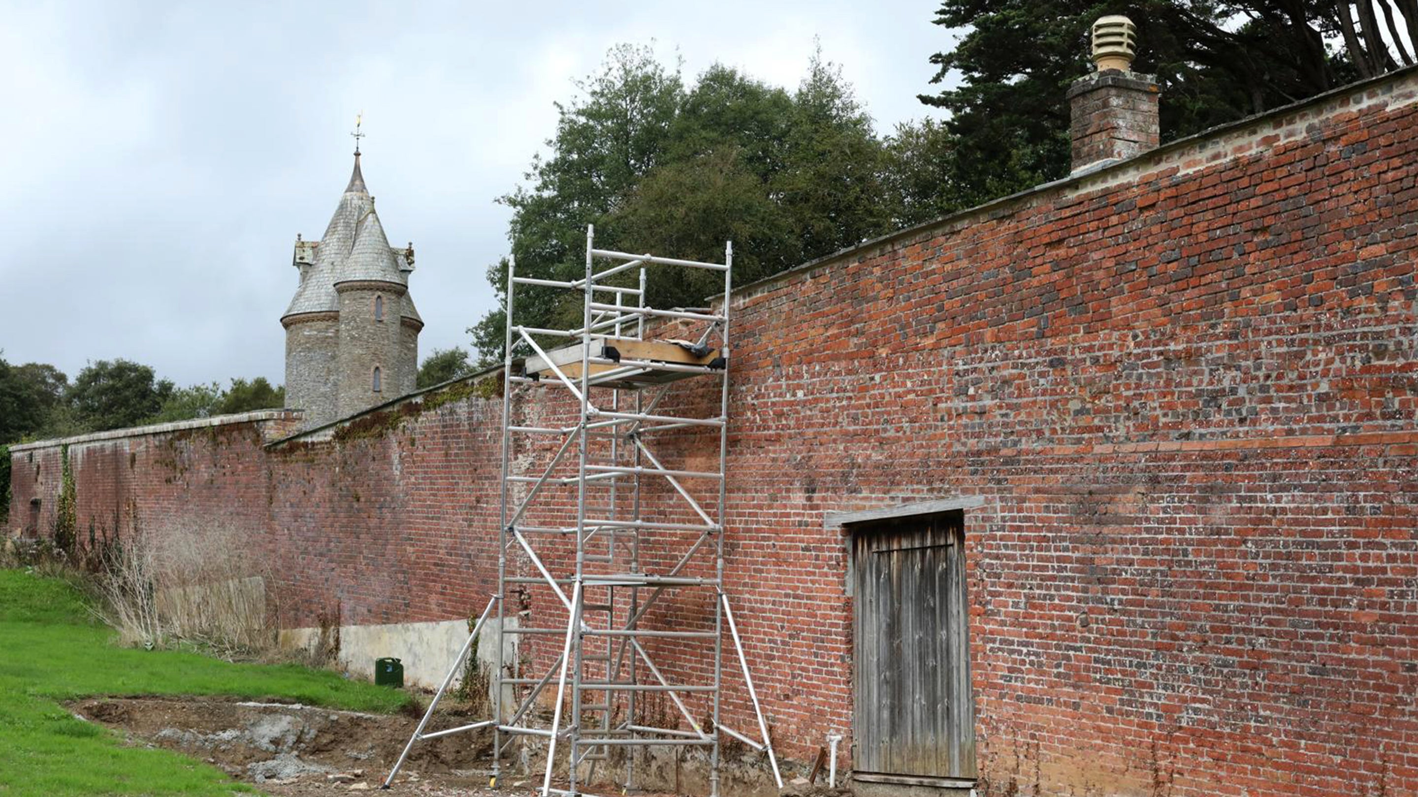 An image showing the first stage of the glasshouse foundations in front of the red brick walls of the Walled Garden. The Water Tower is in the background.