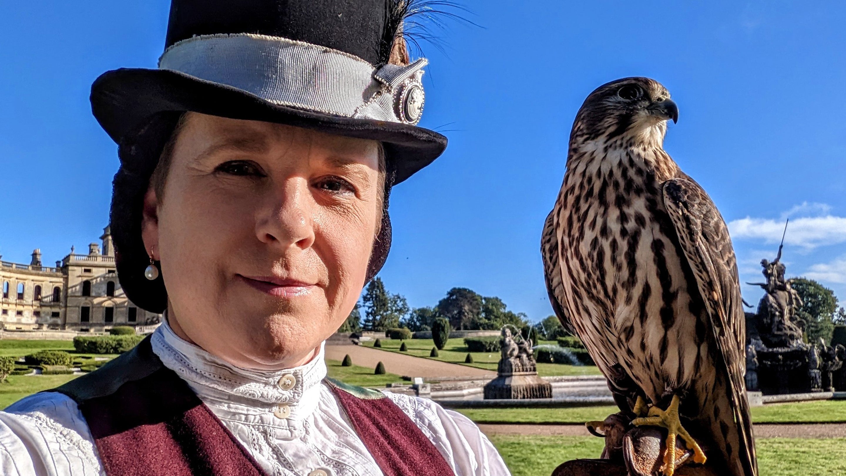The image shows a woman in historical Victorian dress holding a merlin on a sunny day.