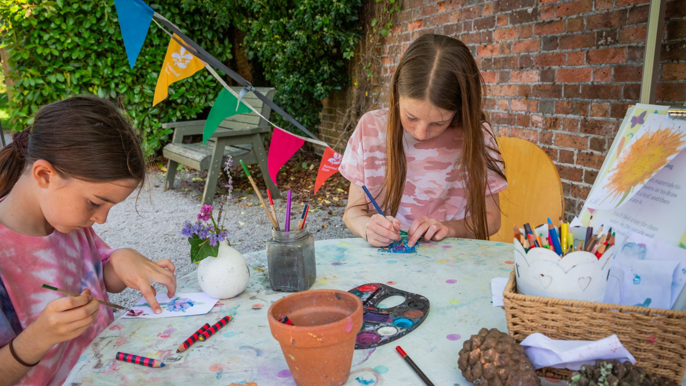 Two girls sat at a crafting table with paints, crayons and paper busy working on their crafts