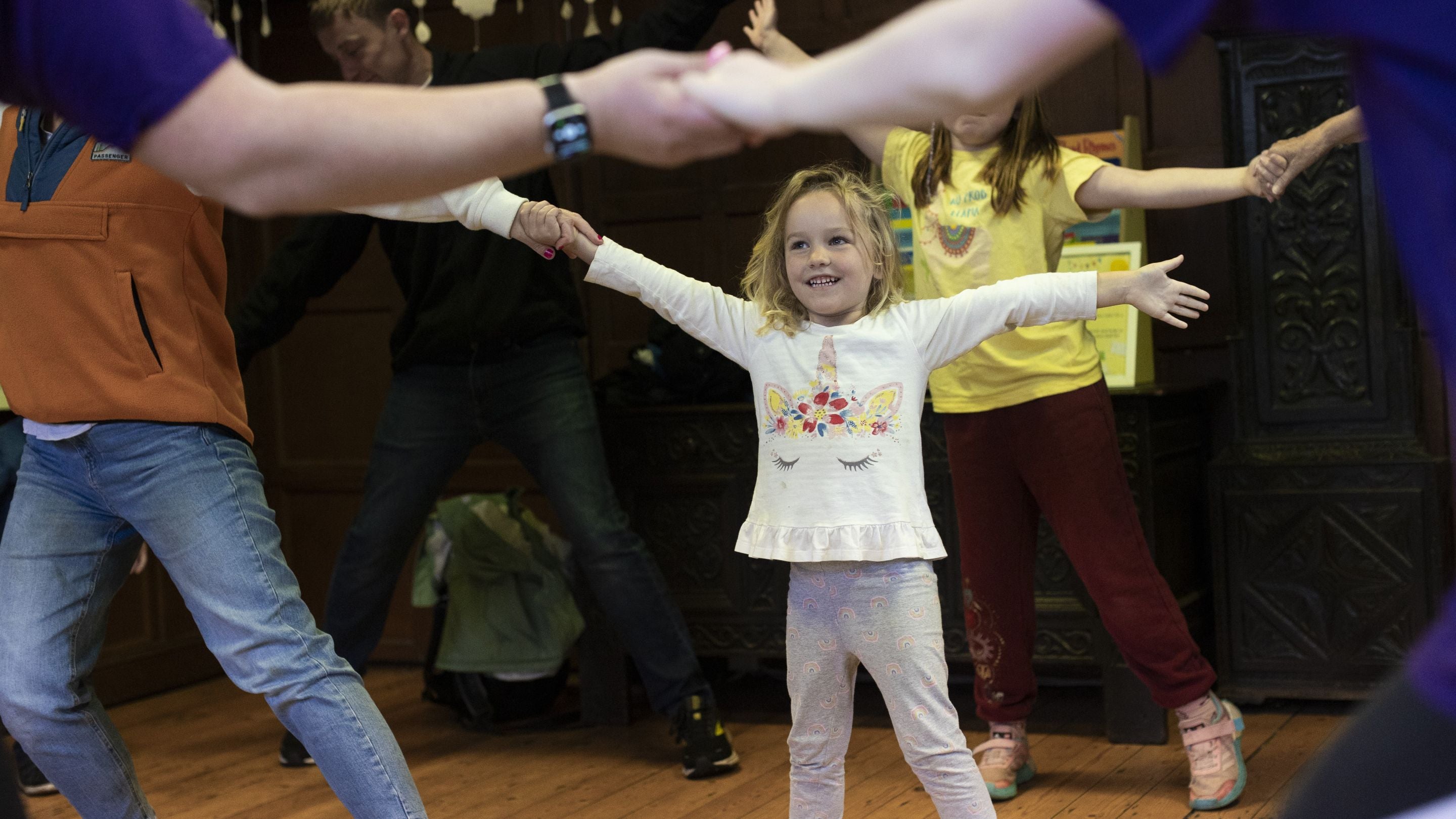 A group of people dancing in a circle, featuring a young girl wearing white in the centre of the group