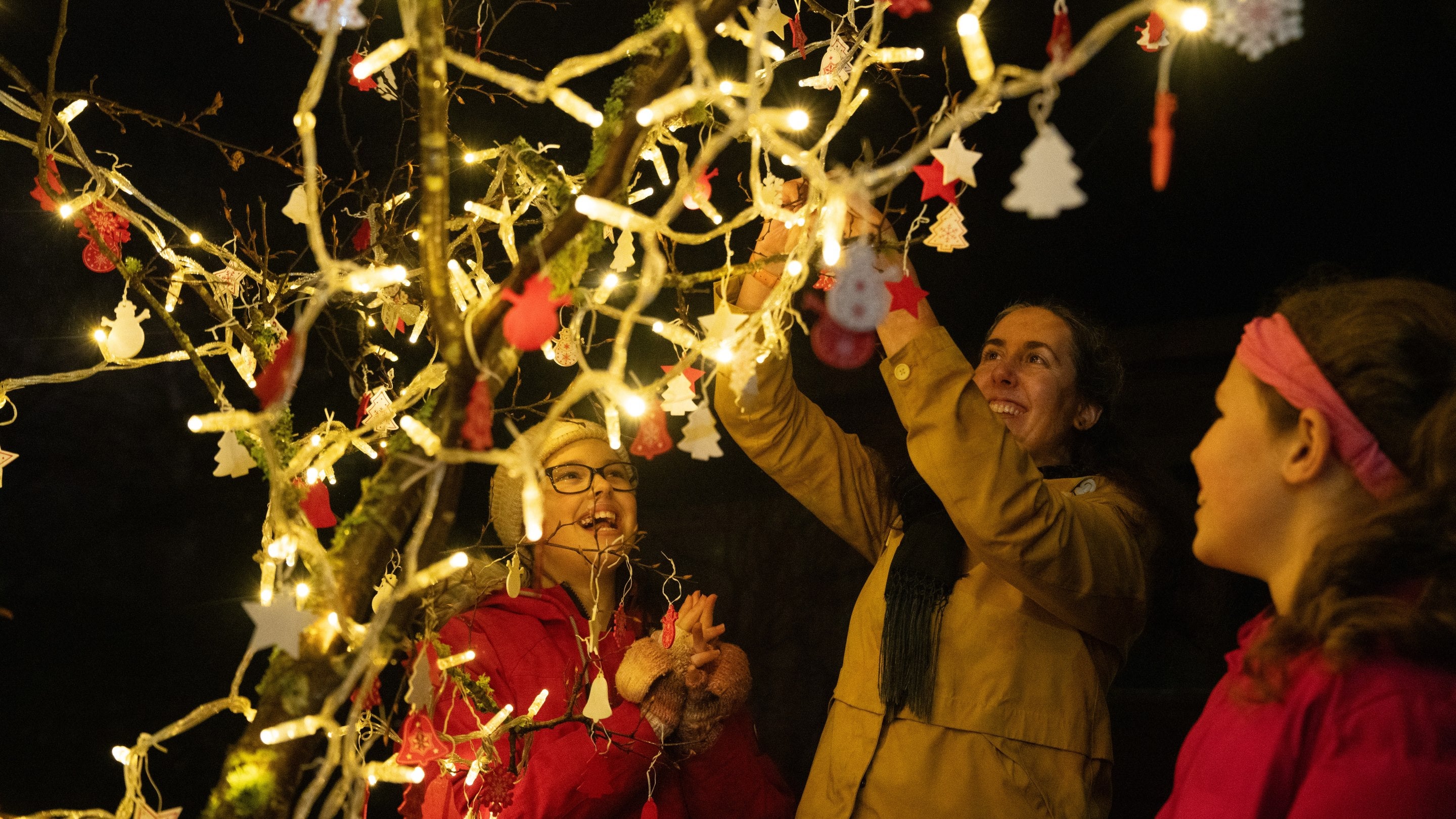 Woman and two children hanging baubles on a tree covered in fairy lights.
