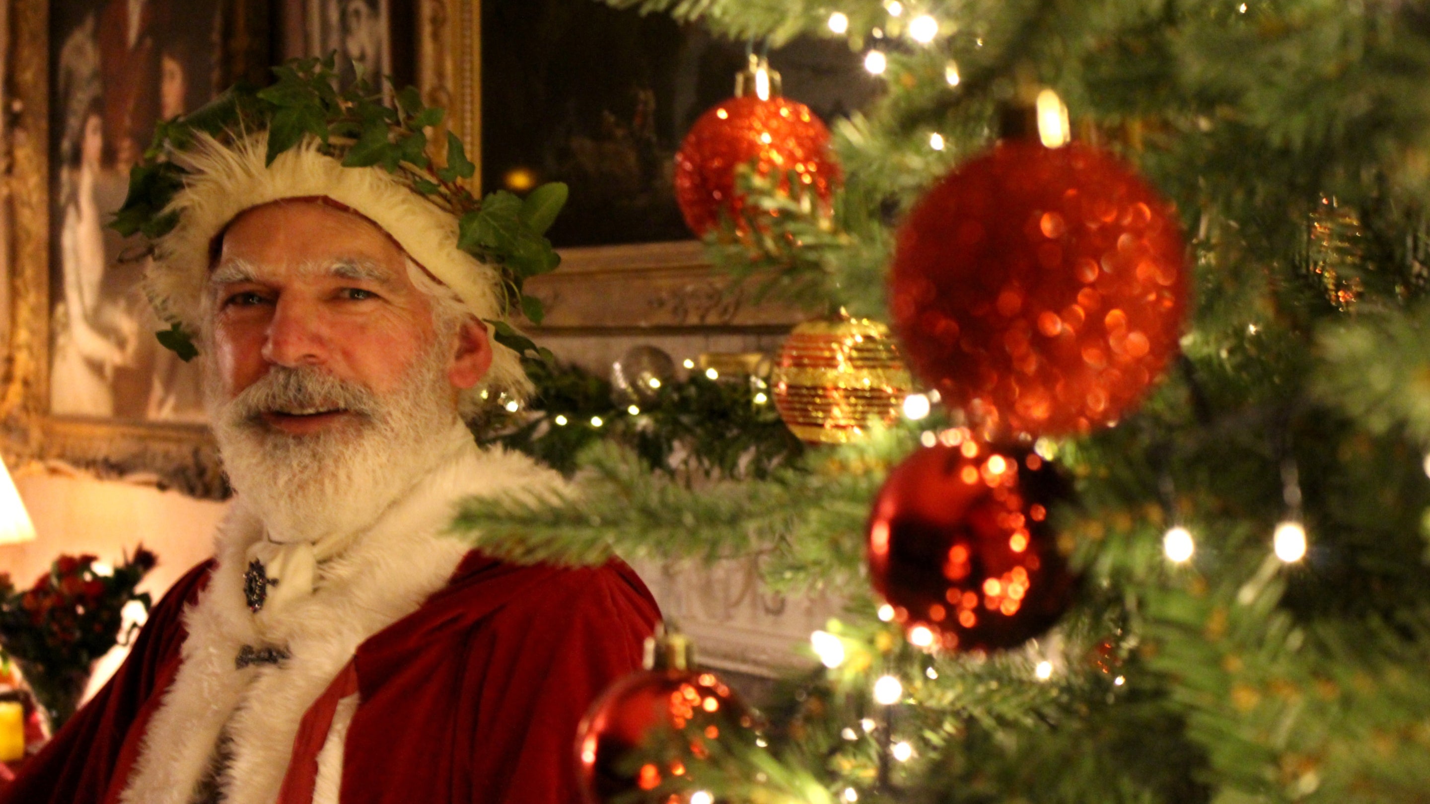 Father Christmas wearing red robes trimmed with white, stood before the fireplace in the drawing room with a Christmas tree in the foreground. The tree is decorated with red and gold baubles, beads and fairy lights. There is a garland on the fireplace with fairy lights.