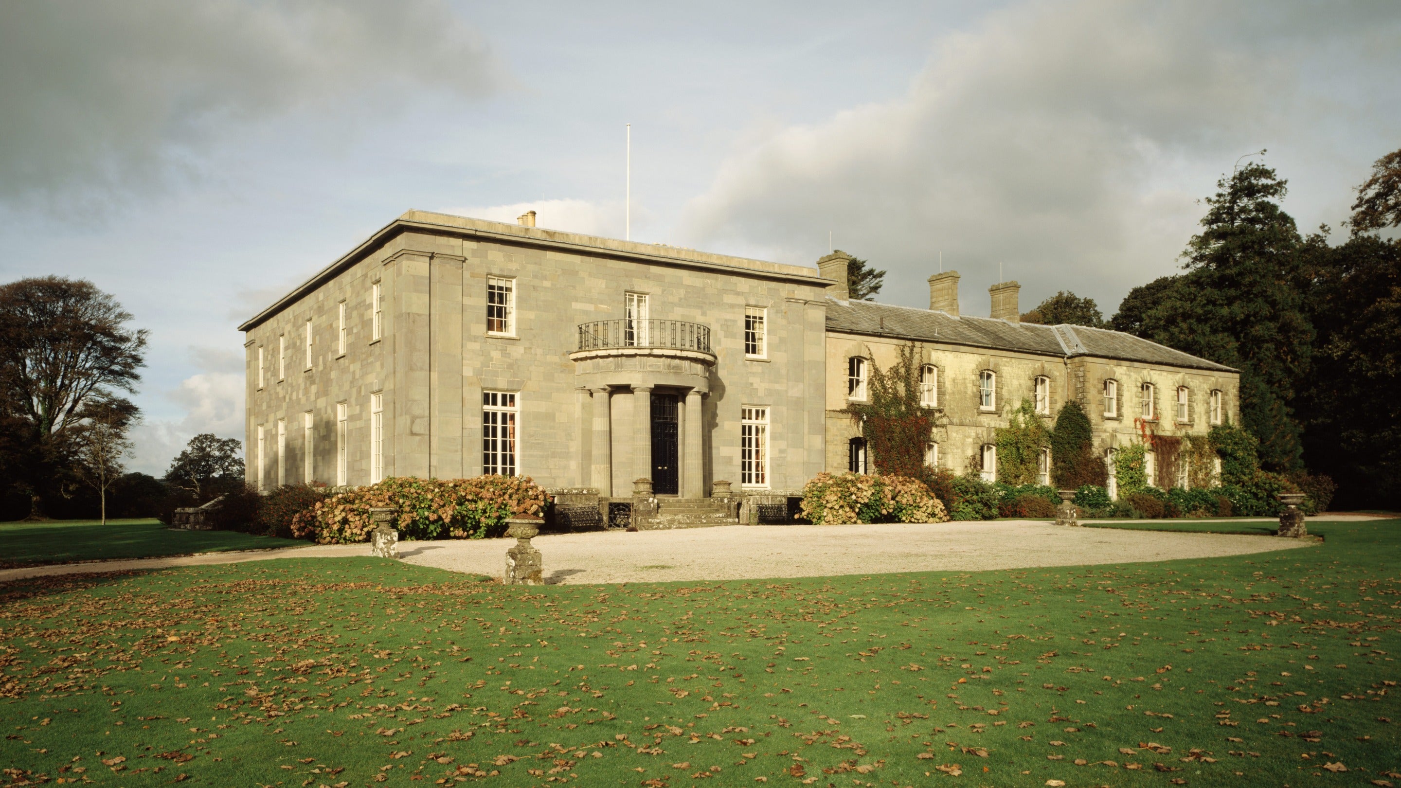 View of the south and east fronts of Arlington Court with semicircular Doric portico and autumn leaves on ground in Devon.