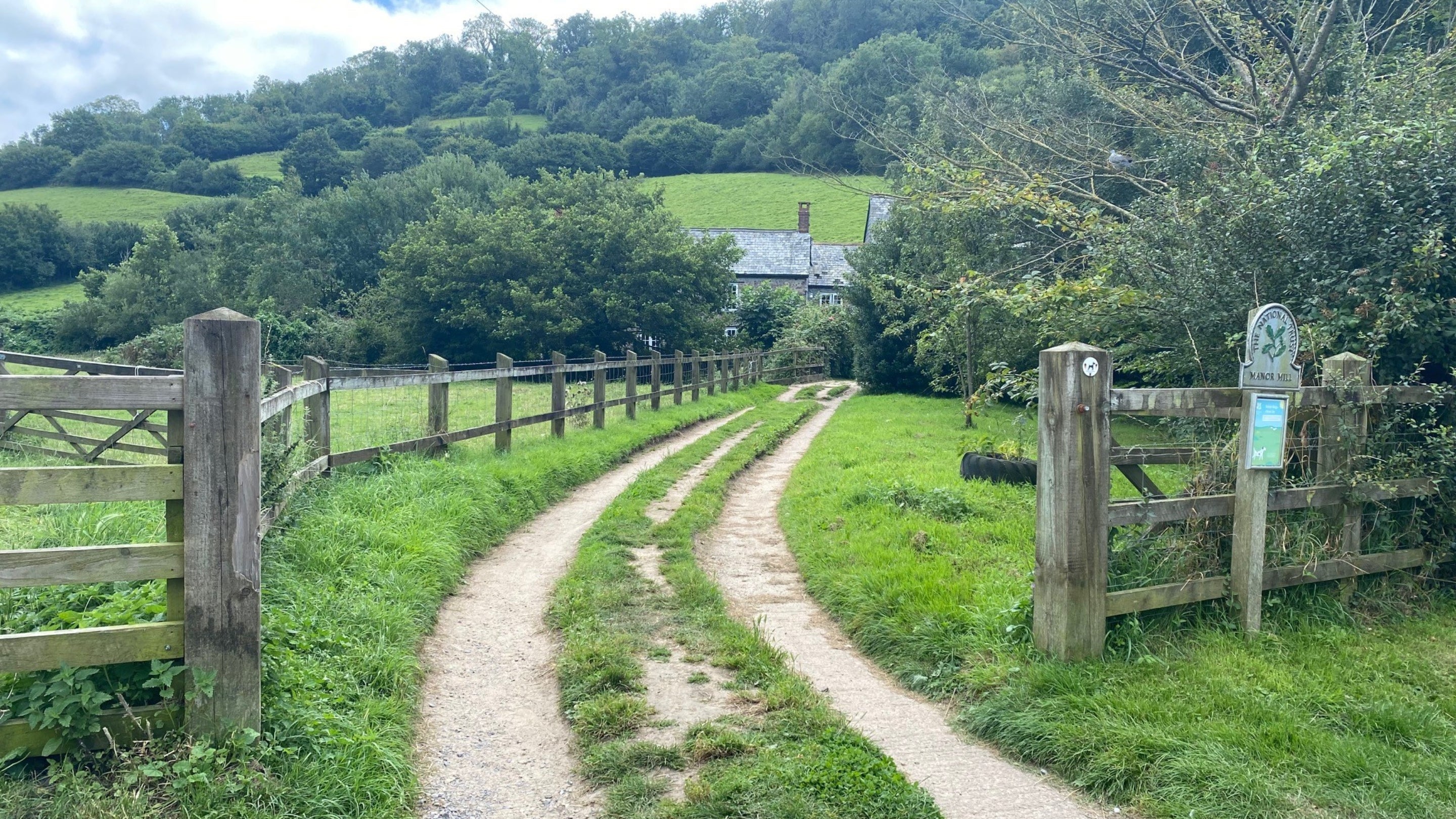 Grassy rutted track with a fence on either side leading to stone buidlings