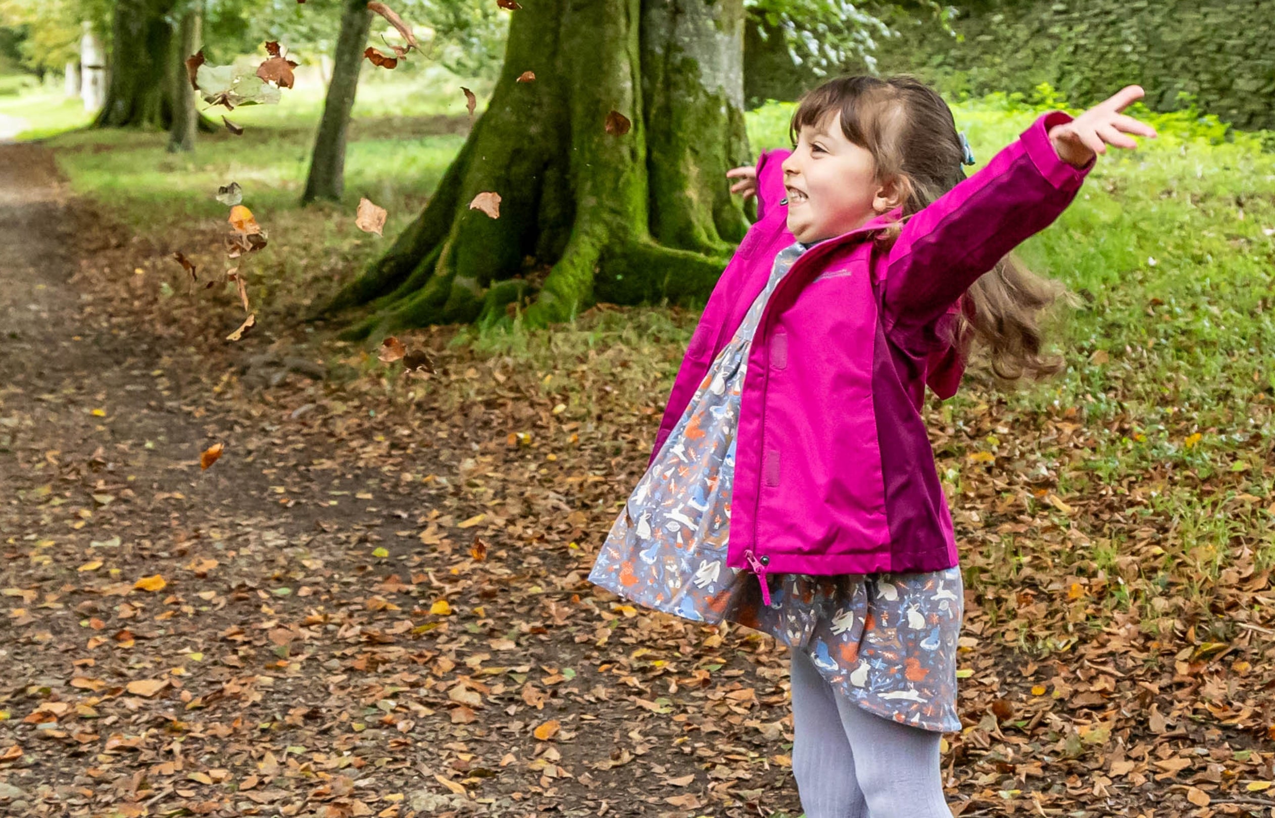 A girl in a pink coat looks happy as she throws fallen leaves into the air