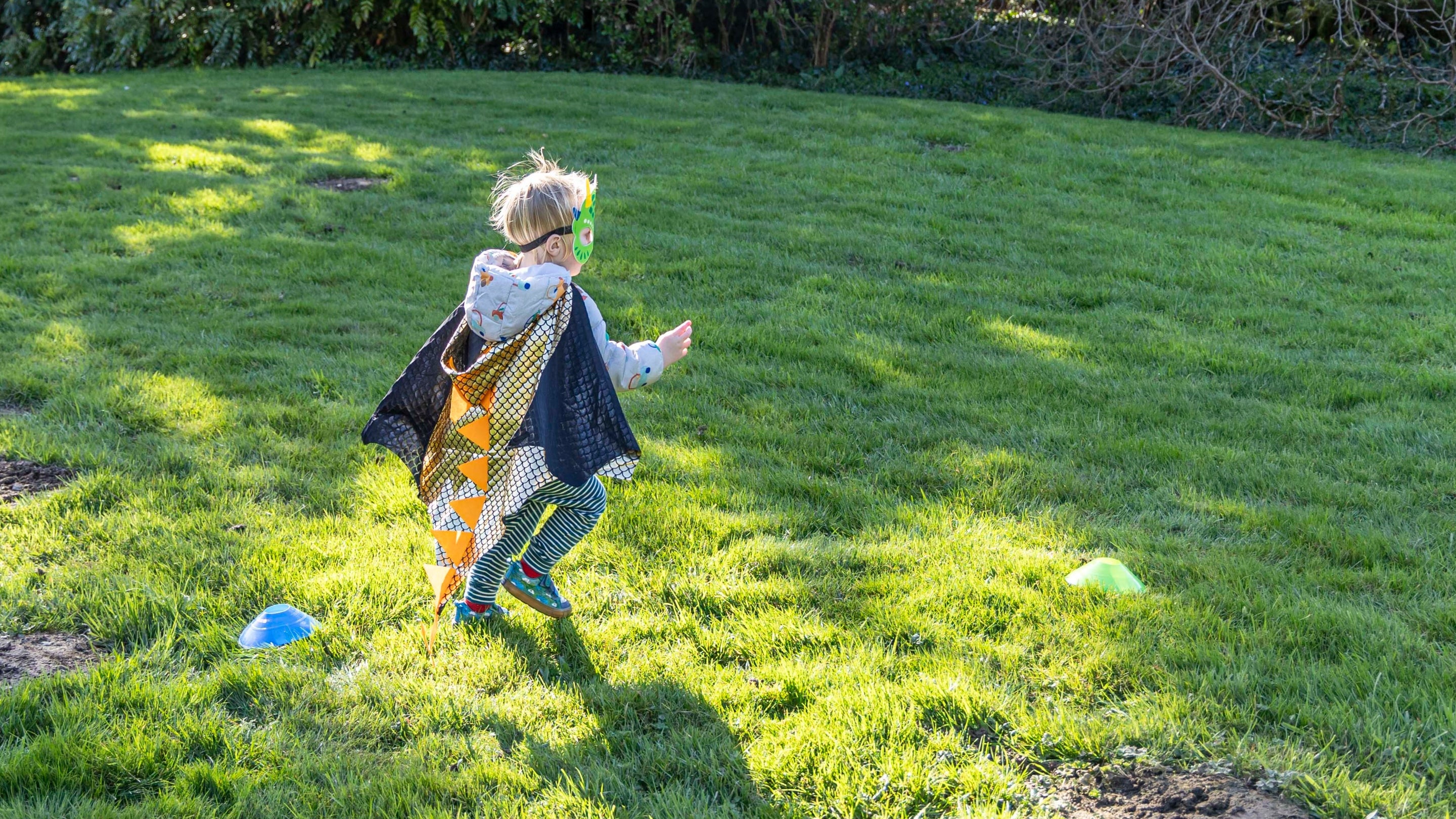A child dressed in a dragon mask and gold cape runs across the grass, facing away from the camera.