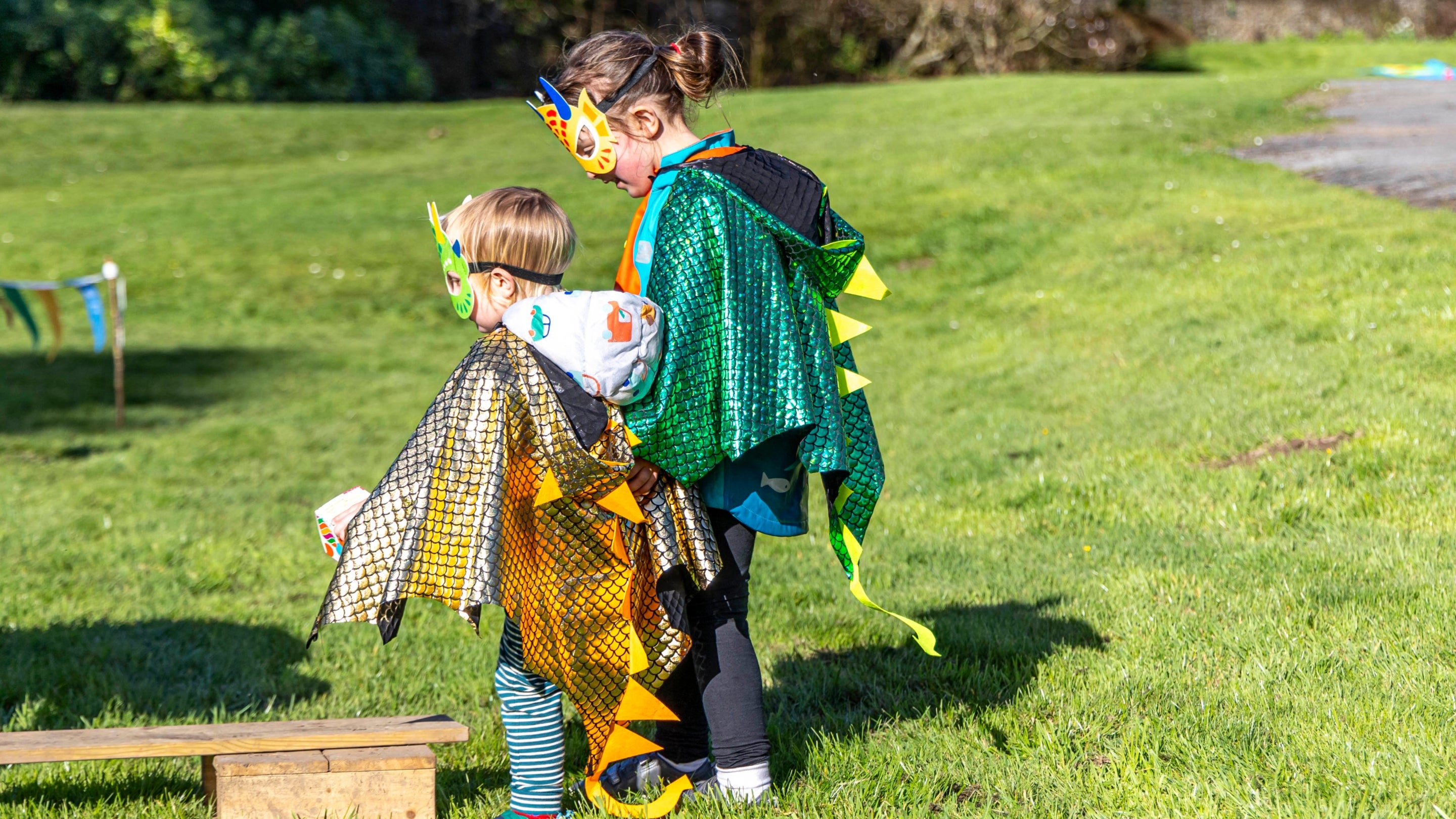 Two children dressed in shiny dragon capes and masks play on the grass.