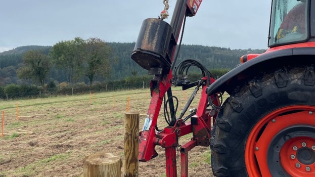 The rear wheel of an orange tractor is visible next to a wooden post, with a large metal post basher attached. Views across a field with trees in the distance are visible behind.