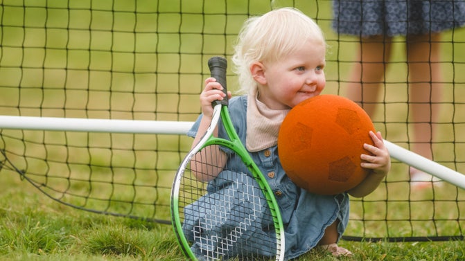 A blonde toddler wearing a blue dress sits in front of a tennis net, holding a green tennis racquet and an orange football