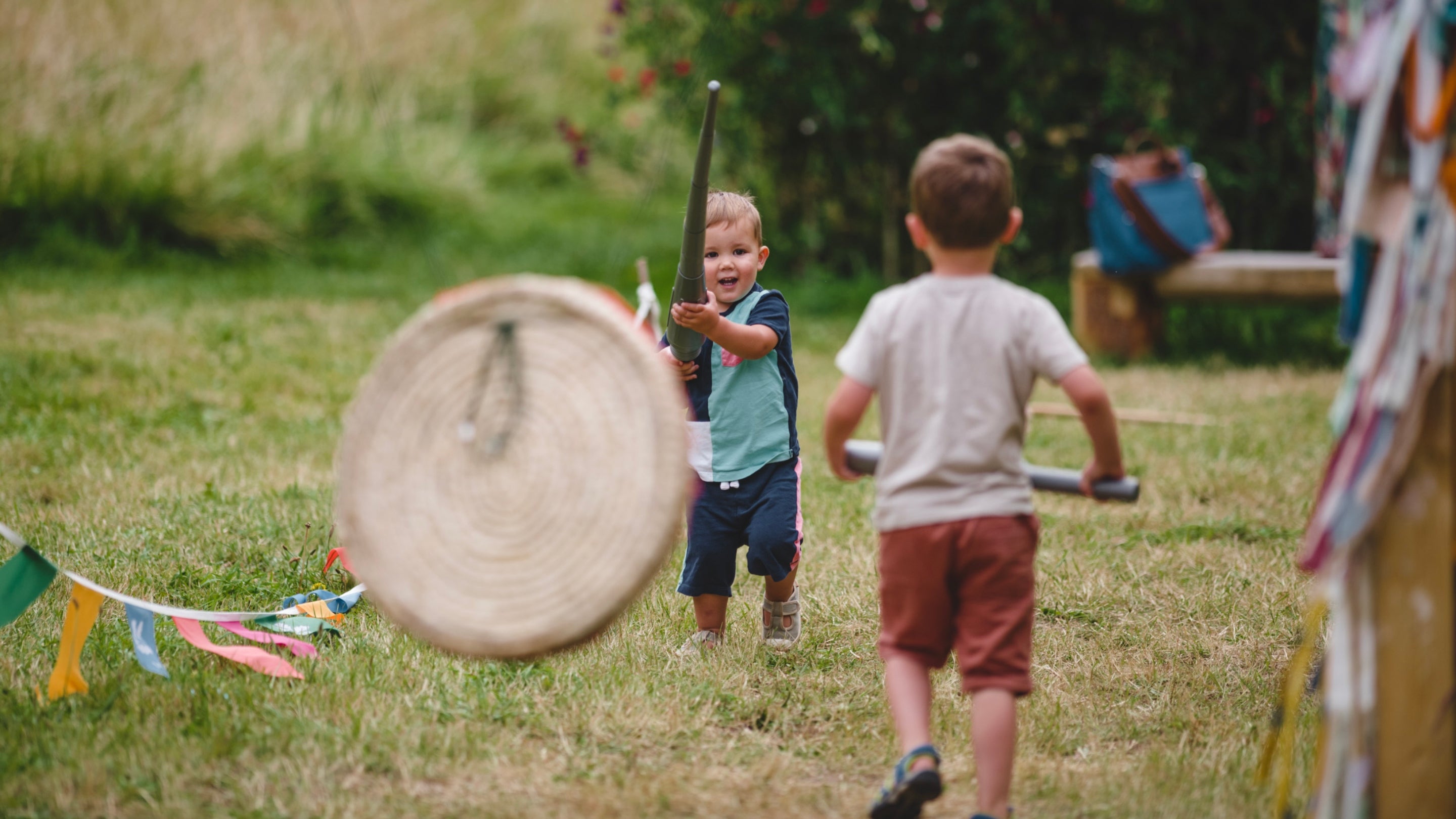 A young boy stands in the centre of the image with a toy lance. Another child is facing towards him, slightly out of focus, in the foreground, and colourful flags run alongside the children