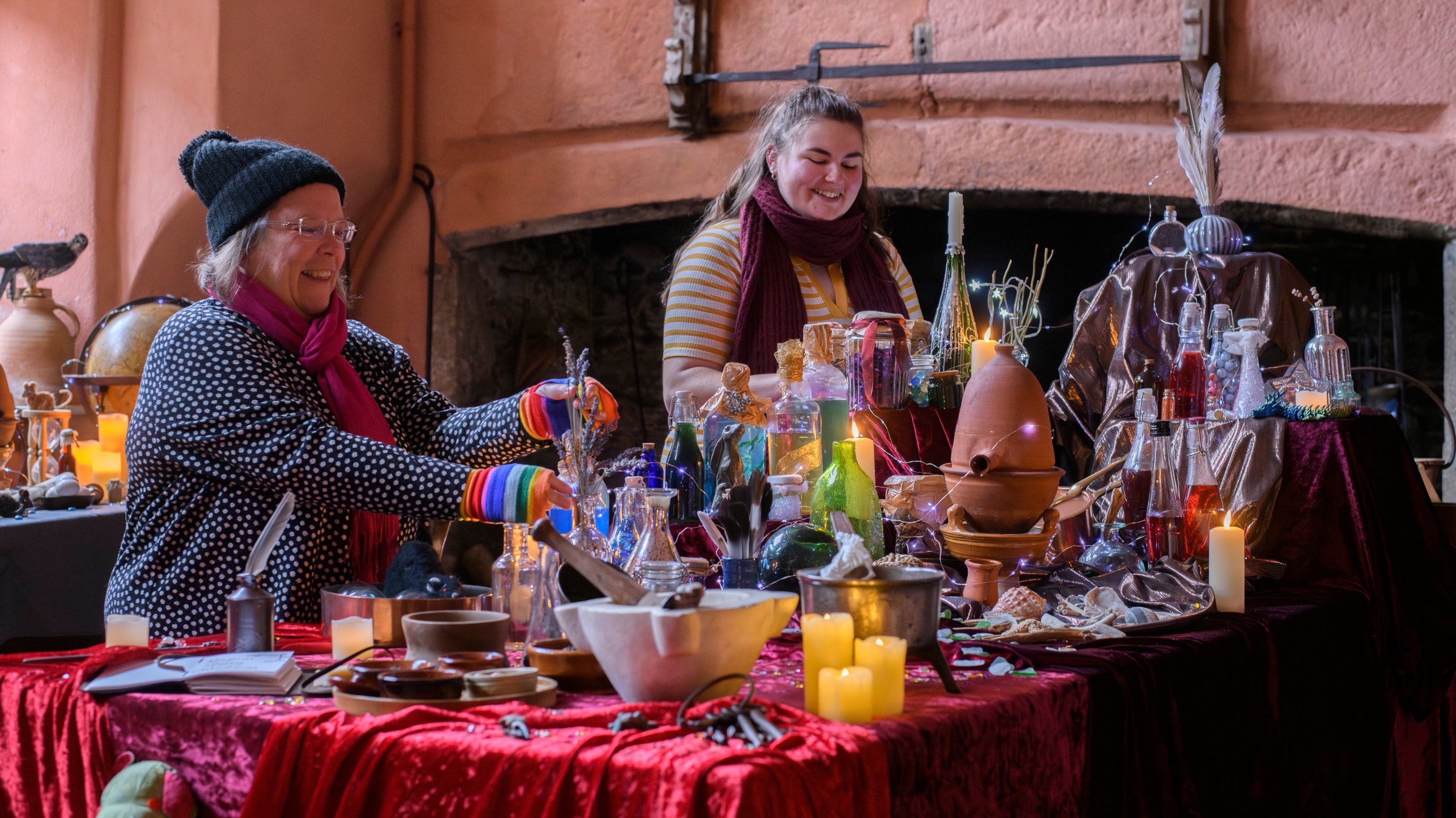 A table covered with coloured potions in glass bottles, fairy lights and earthenware bowls is in the foreground, with two women dressed in winter clothing behind.