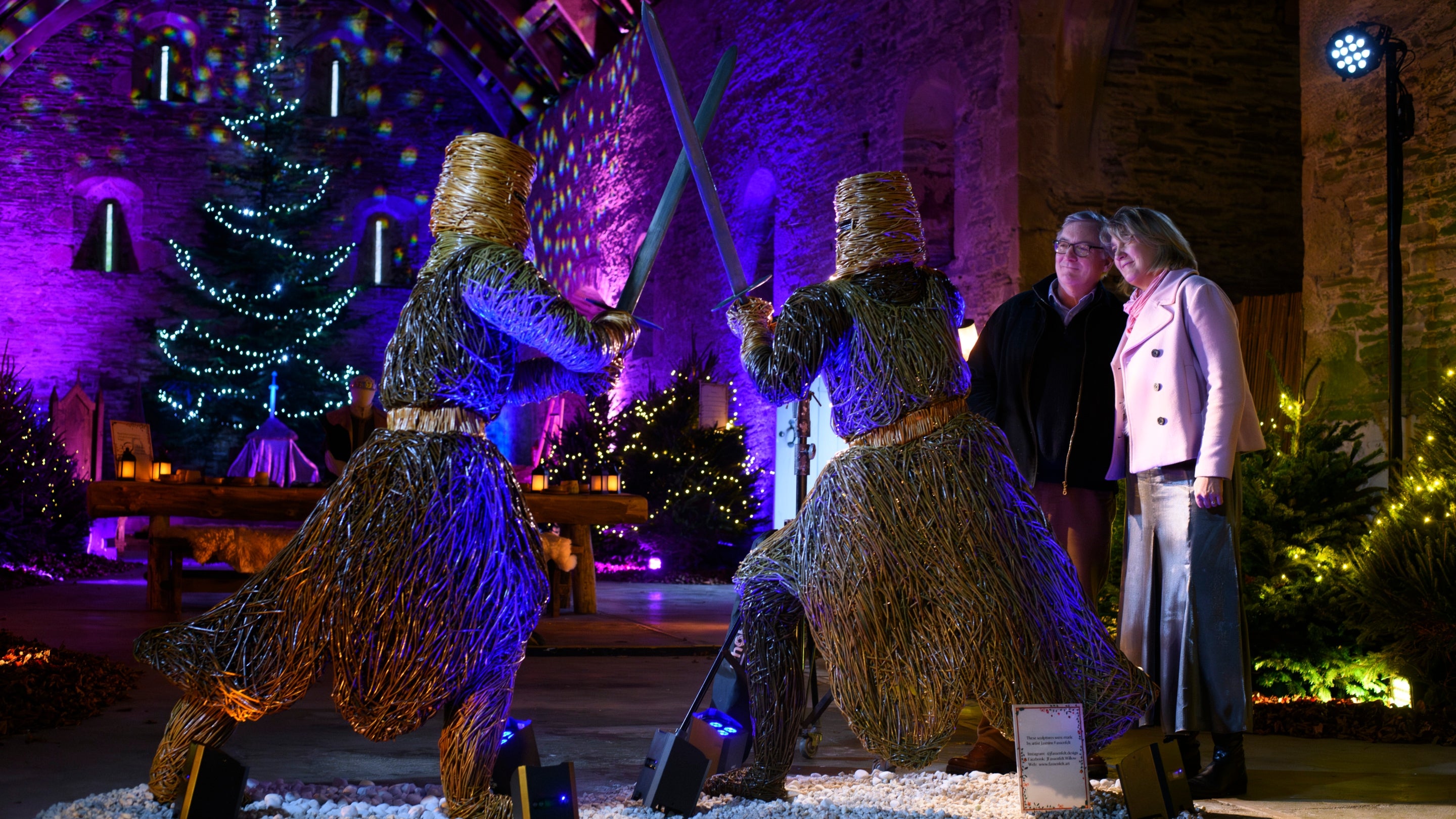 Two visitors admire a life sized willow sculpture of two knights duelling. There are Christmas trees with white fairy lights in the background, and the lighting is purple and blue.