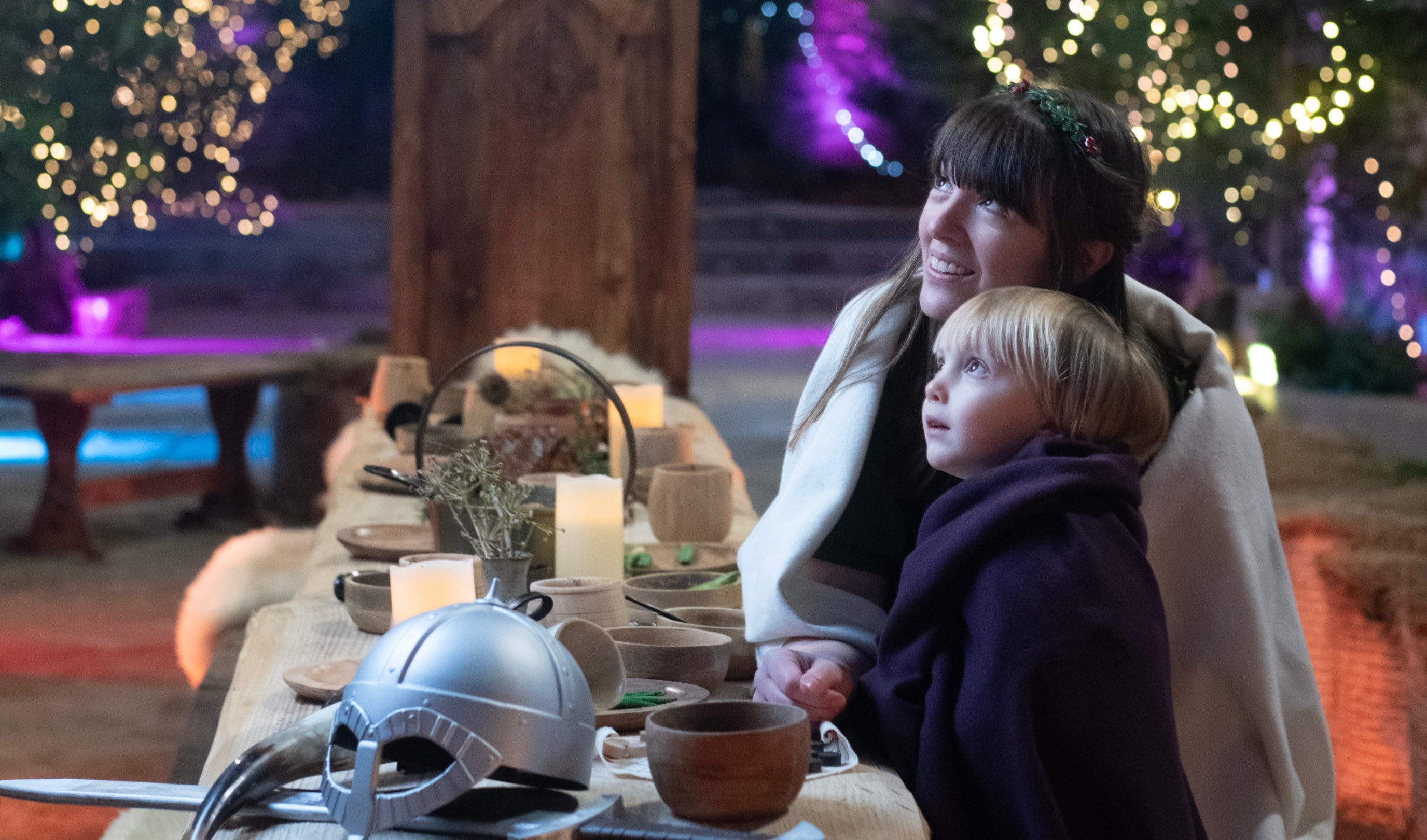 A woman and child wrapped in cloaks sit and look up towards the top left of the shot. They are at a long wooden table covered in wooden cups and bowls with a silver helmet in the foreground.