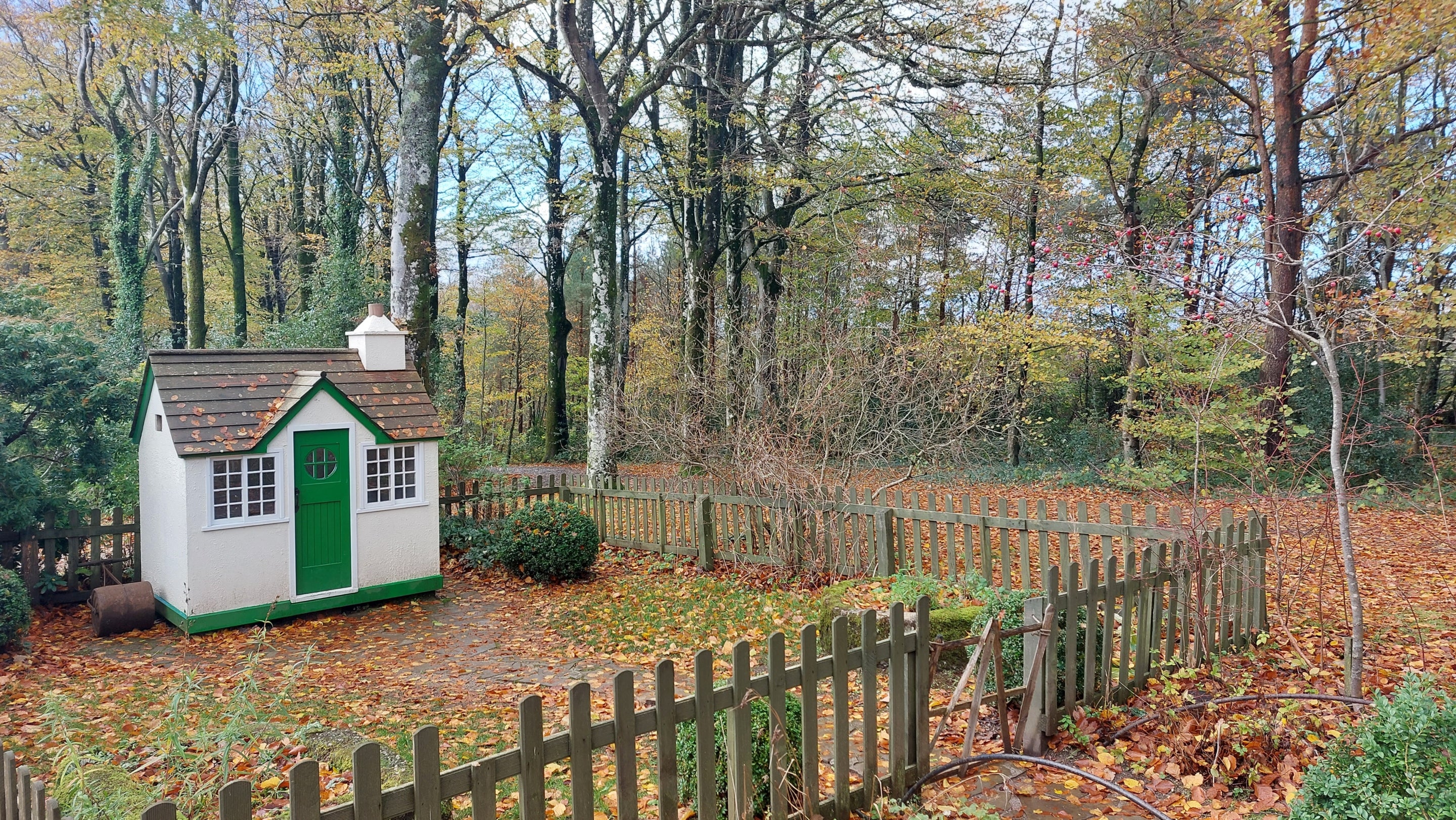 Children's play house with white walls and a green door. There is a small garden in front of the house and a fence.