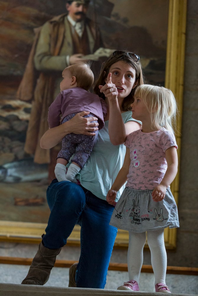 A mum and two kids stand at the top of the central staircase looking down towards the camera with the portrait of Julius Drewe behind them.