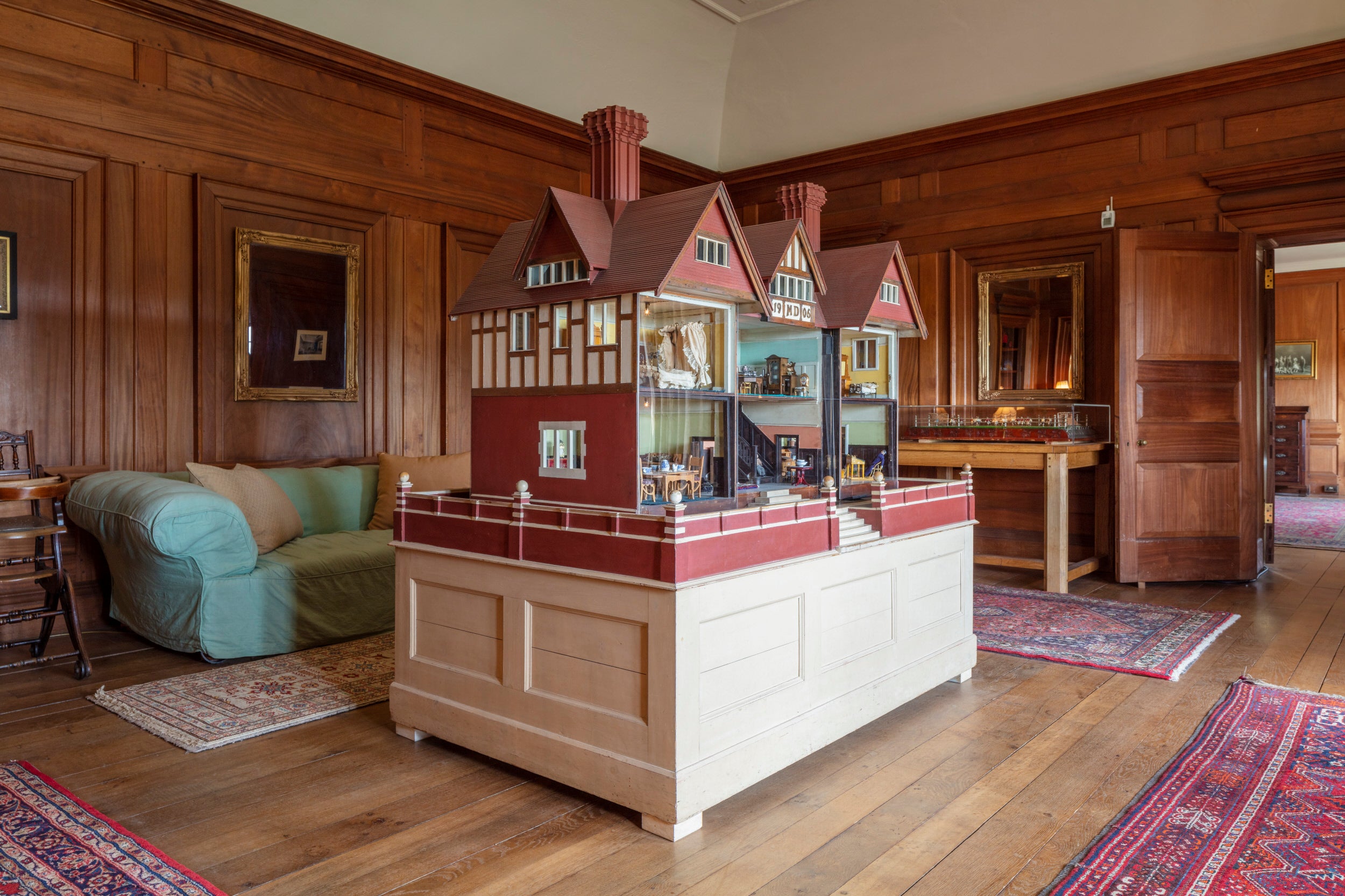 A wood panelled room with a large wooden dolls house in the centre there are rugs on the floor and a green sofa behind the dolls house