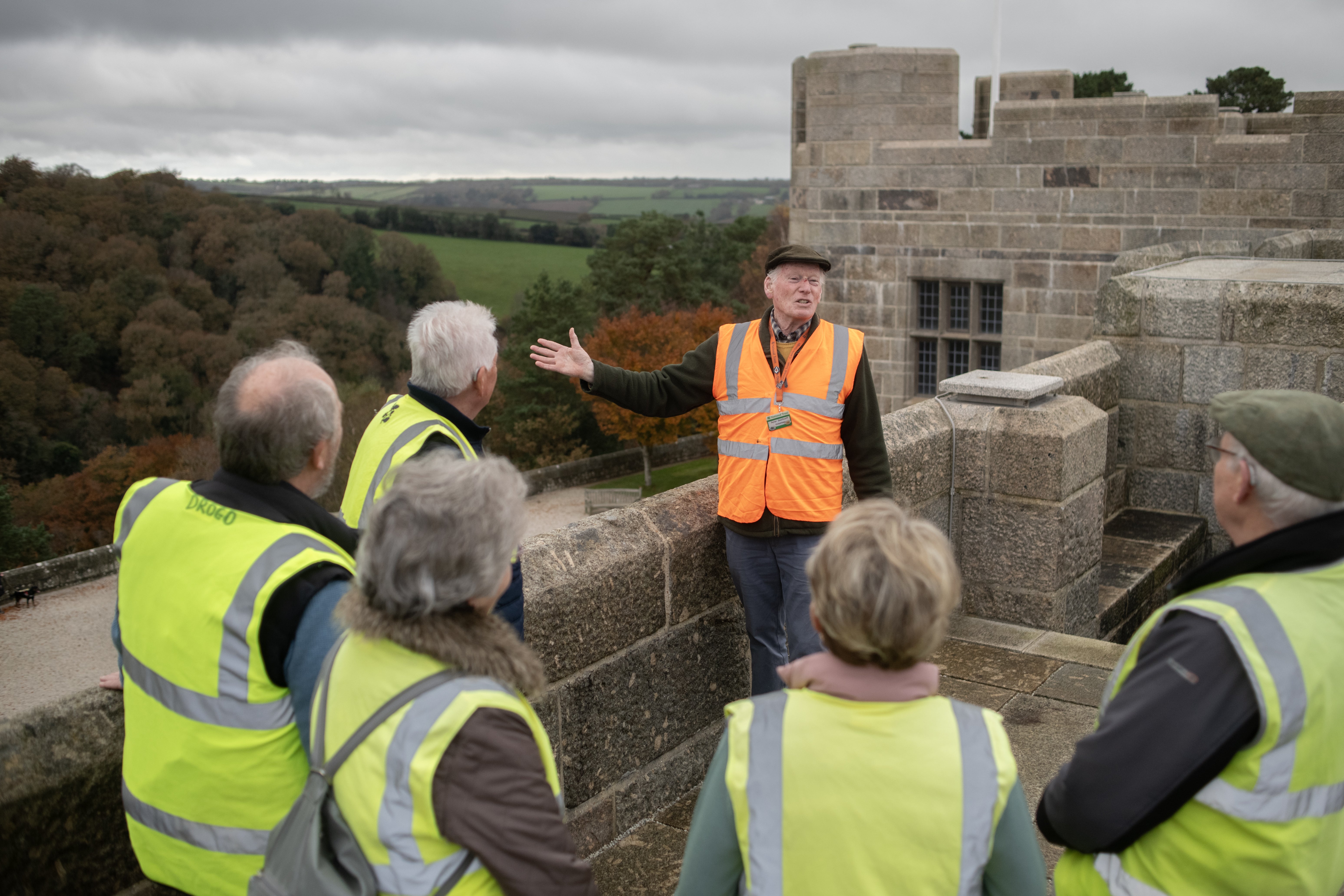 A guide in an orange hi-vis vest shows a group of visitors in yellow hi-vis around the roof at Castle Drogo, while explaining about the landscape and construction work.