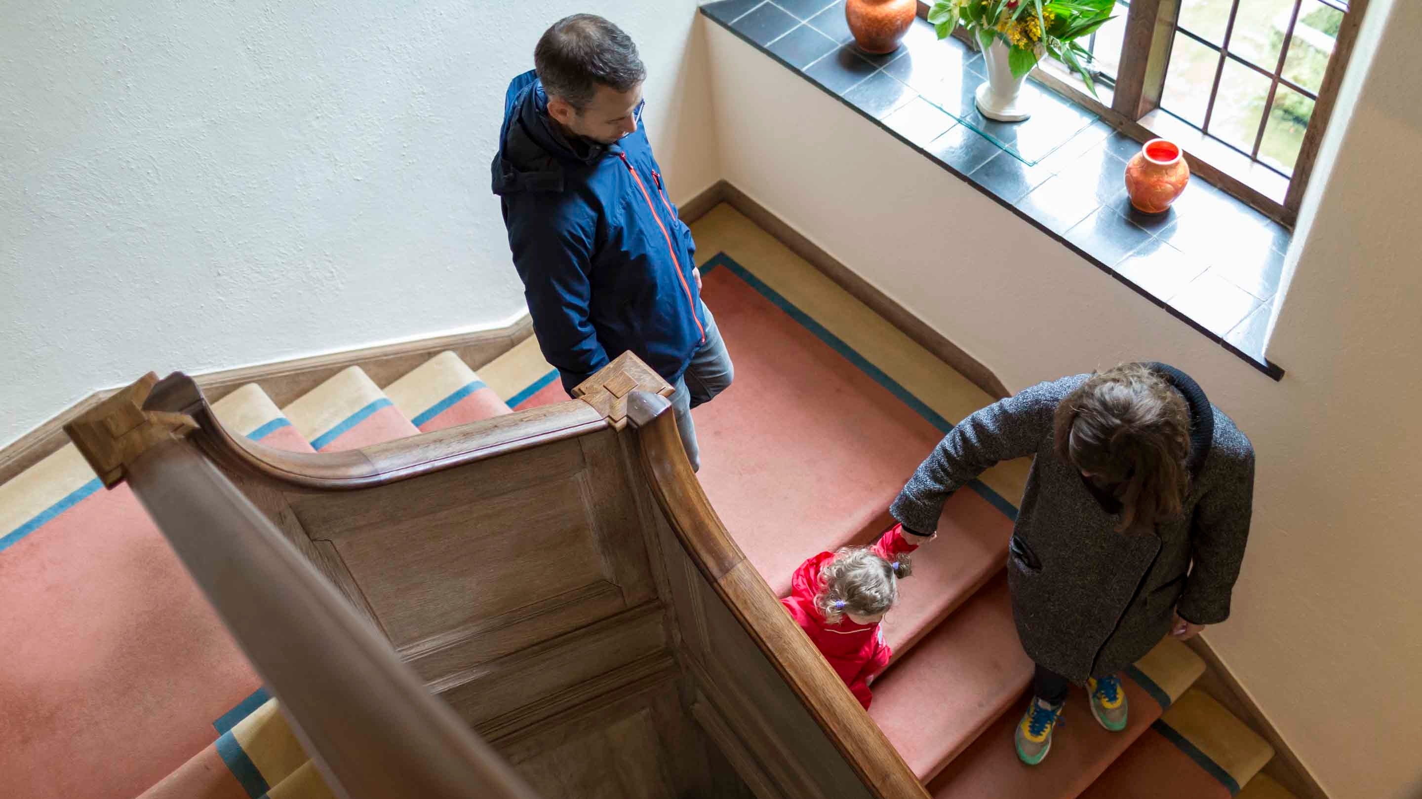 Two adults and a child walking down a stairwell with wooden banisters and red carpet at Coleton Fishacre.