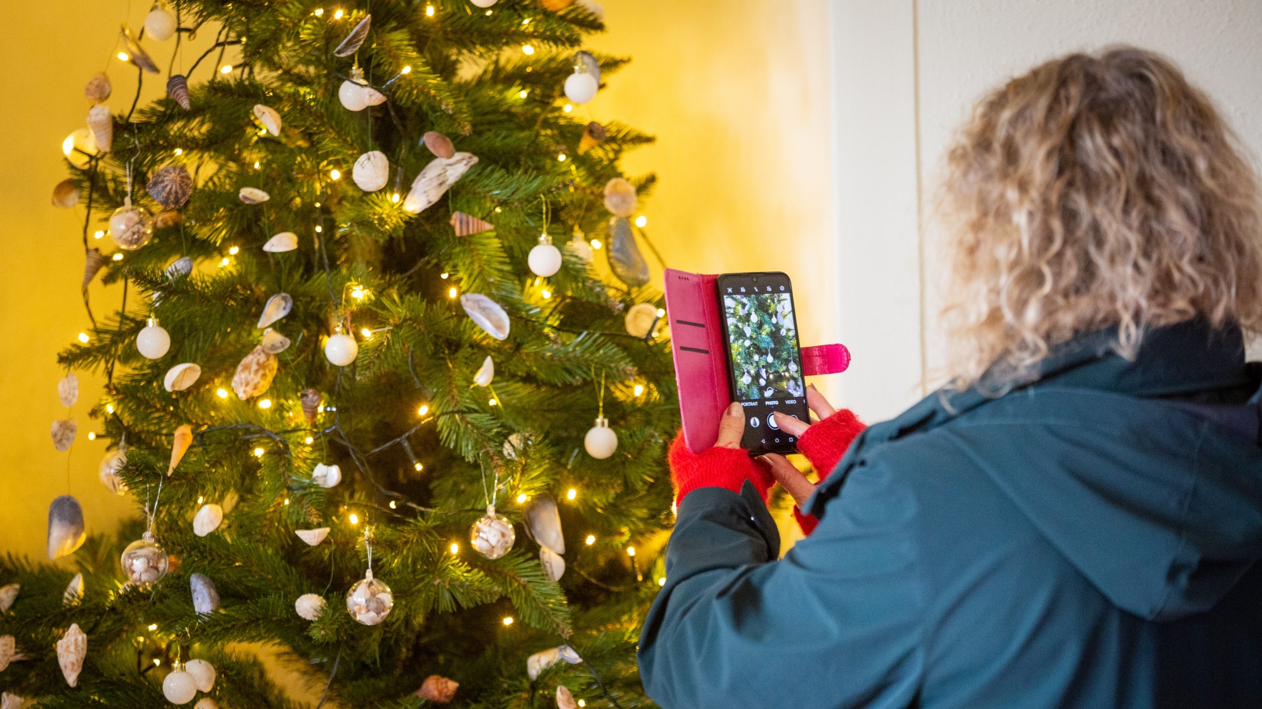 A visitor taking a photo on their phone of a Christmas tree decorated with shell ornaments.