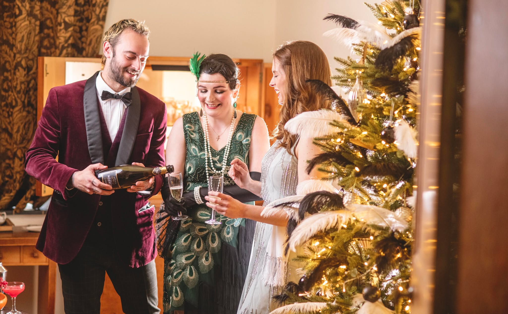 A festive scene with three people in vintage attire. A man in a velvet jacket pours champagne for two women near a decorated, illuminated tree.