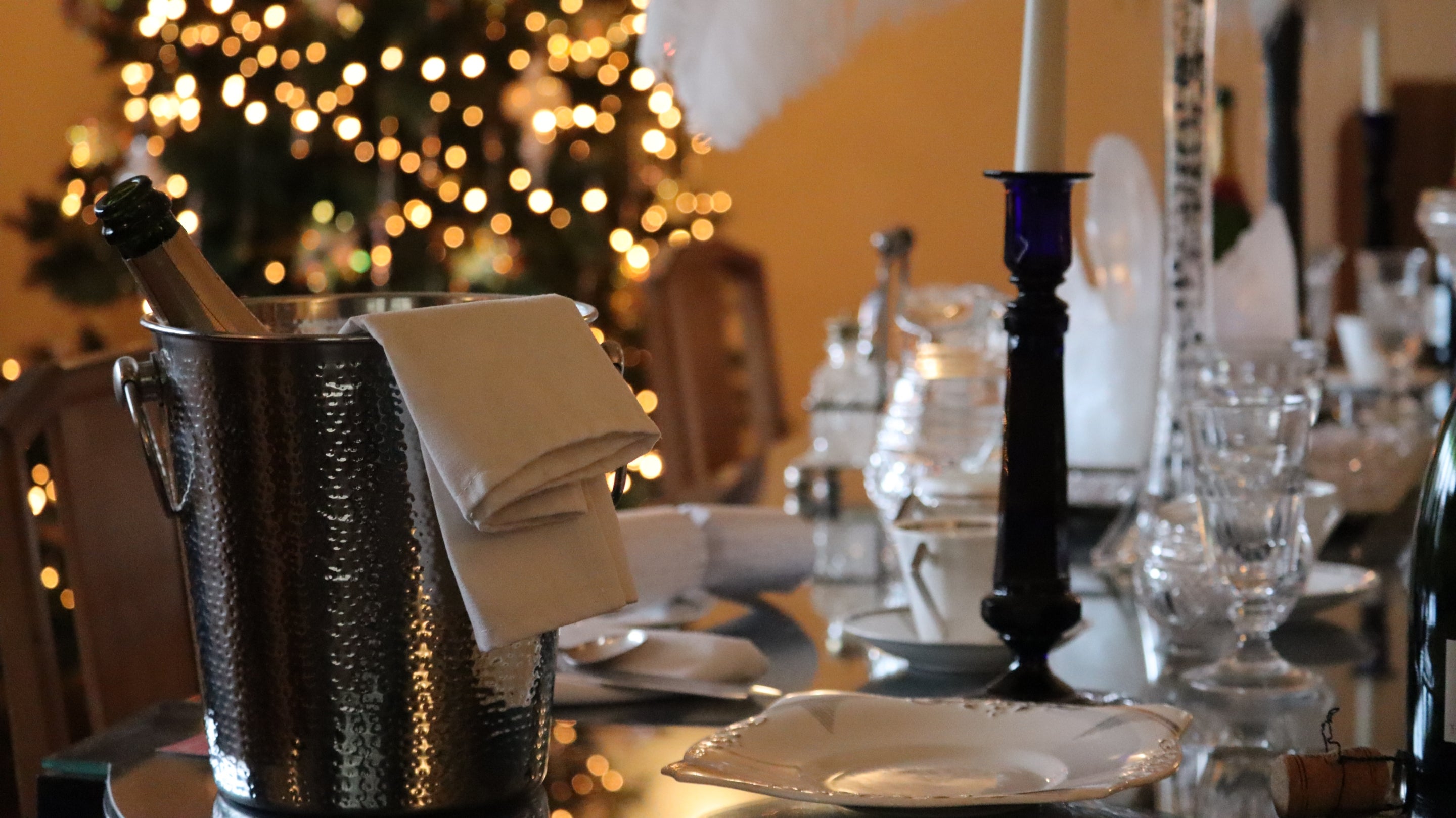 Silver champagne bucket and bottle sits on a table dressed for dinner, with a glittering decorated Christmas tree in the background.