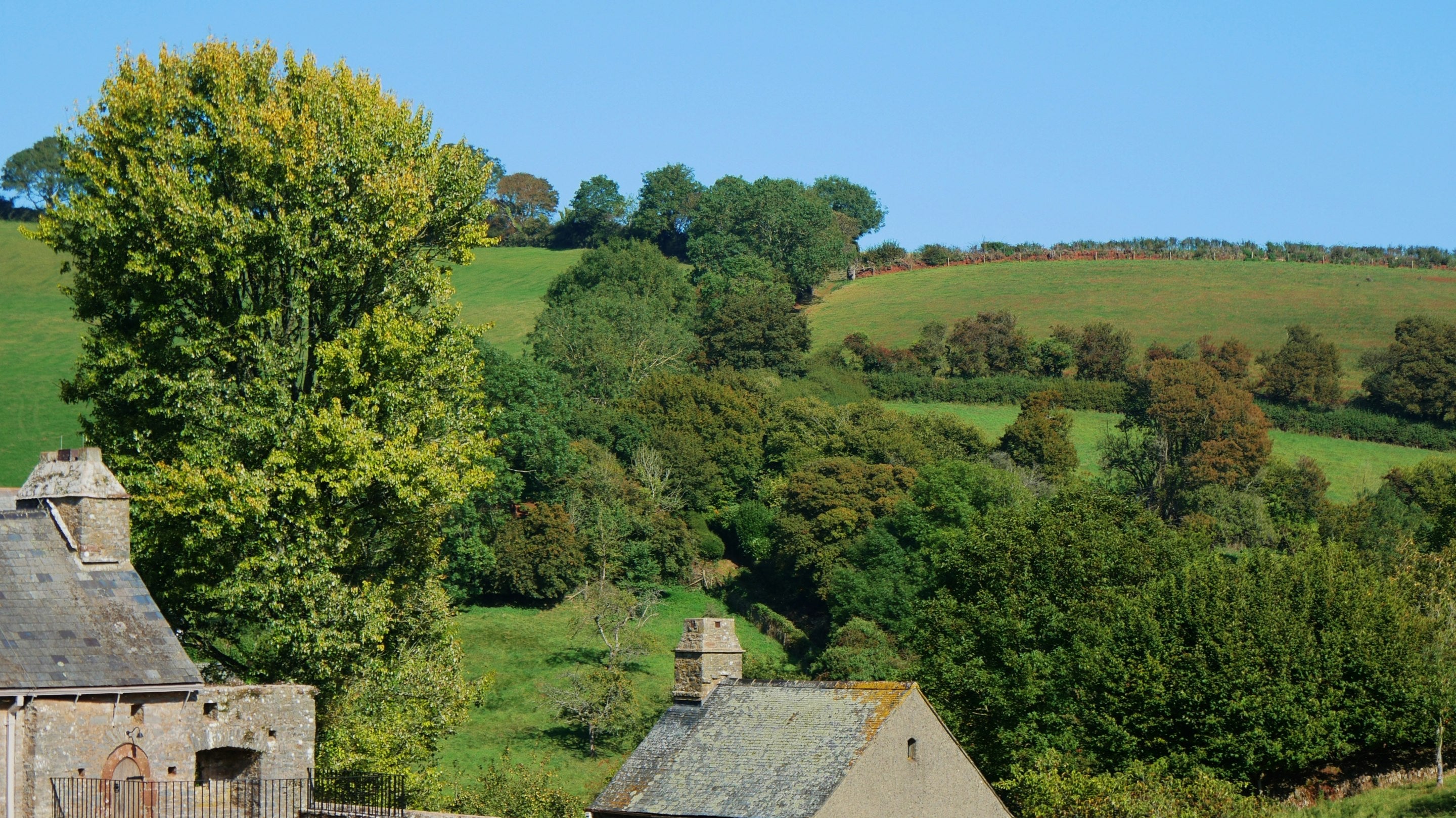 Stone buildings stand in front of woodland and farmed fields in a valley on a sunny day.