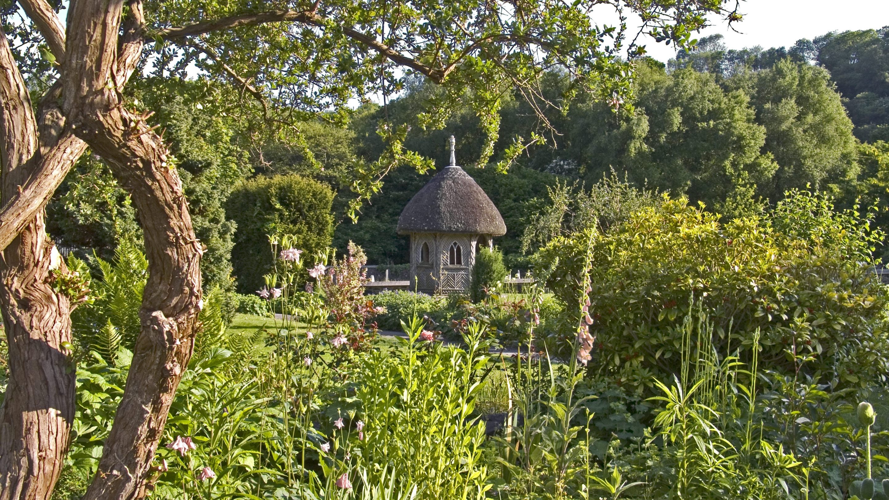 The Garden at Finch Foundry in Devon, a cottage style garden filled with flowers and foliage with a small thatched wooden summerhouse.
