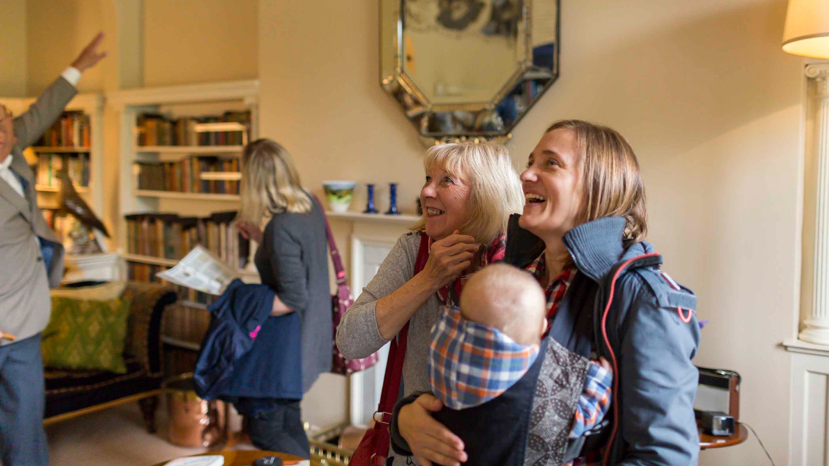 Visitors inside the library at Greenway, Devon. One visitor has a baby in a baby carrier, smiling as they explore the room. In the background a volunteer can be seen pointing out something to a visitor.