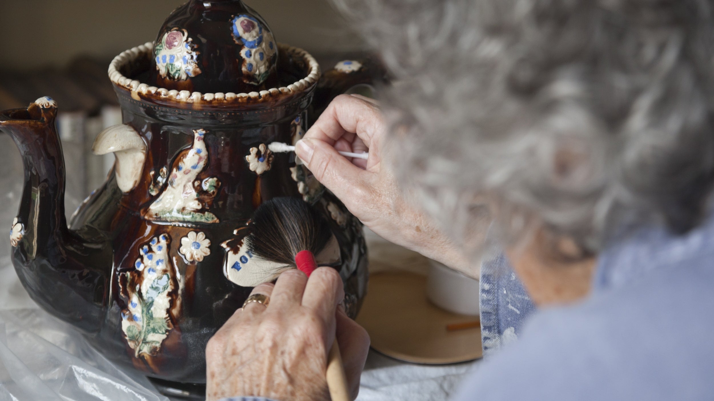 Elderly volunteer carefully cleaning decorative ceramic teapot with a cotton swab