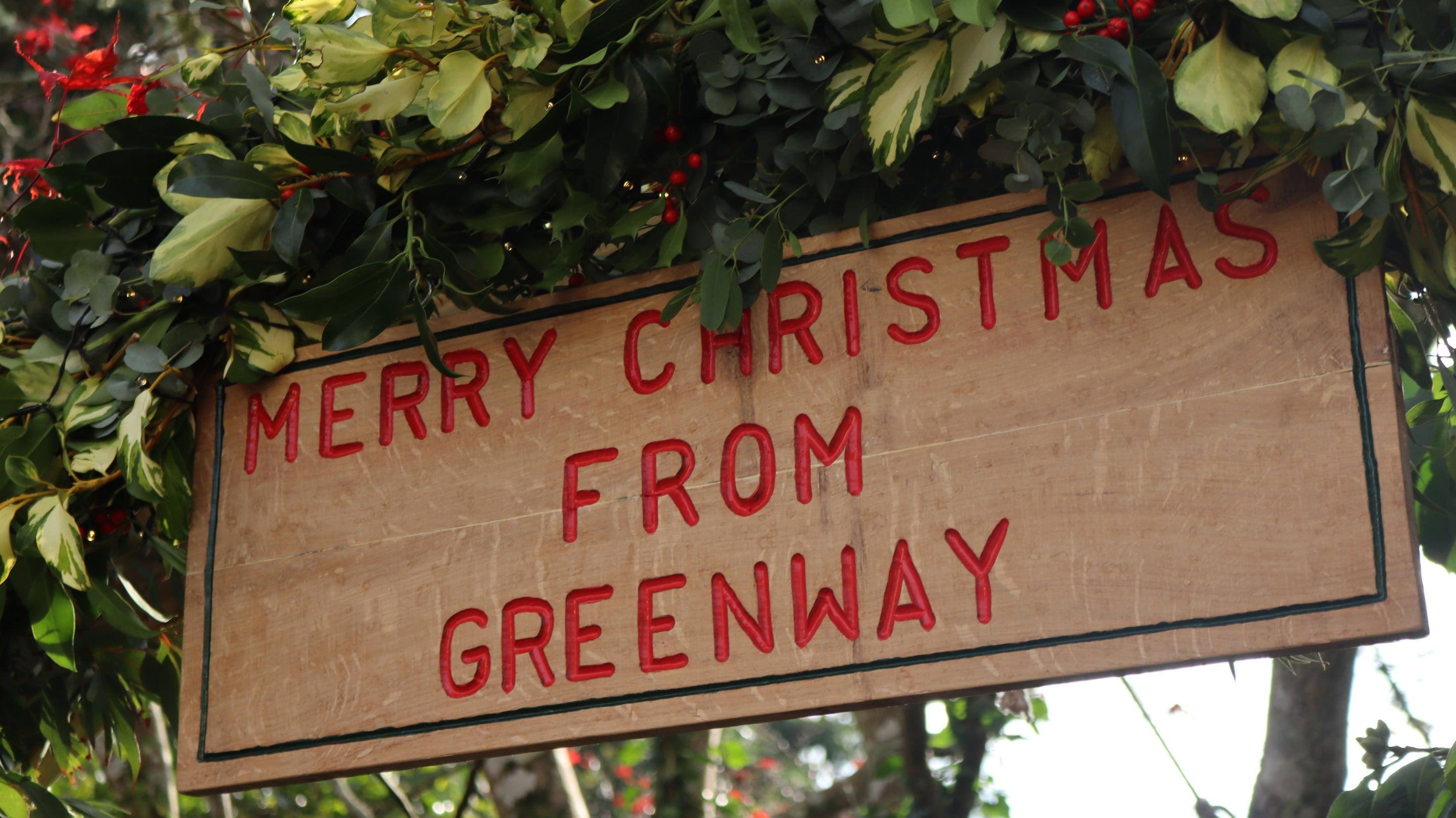 Wooden sign with words Merry Christmas from Greenway with green foliage and red berries.