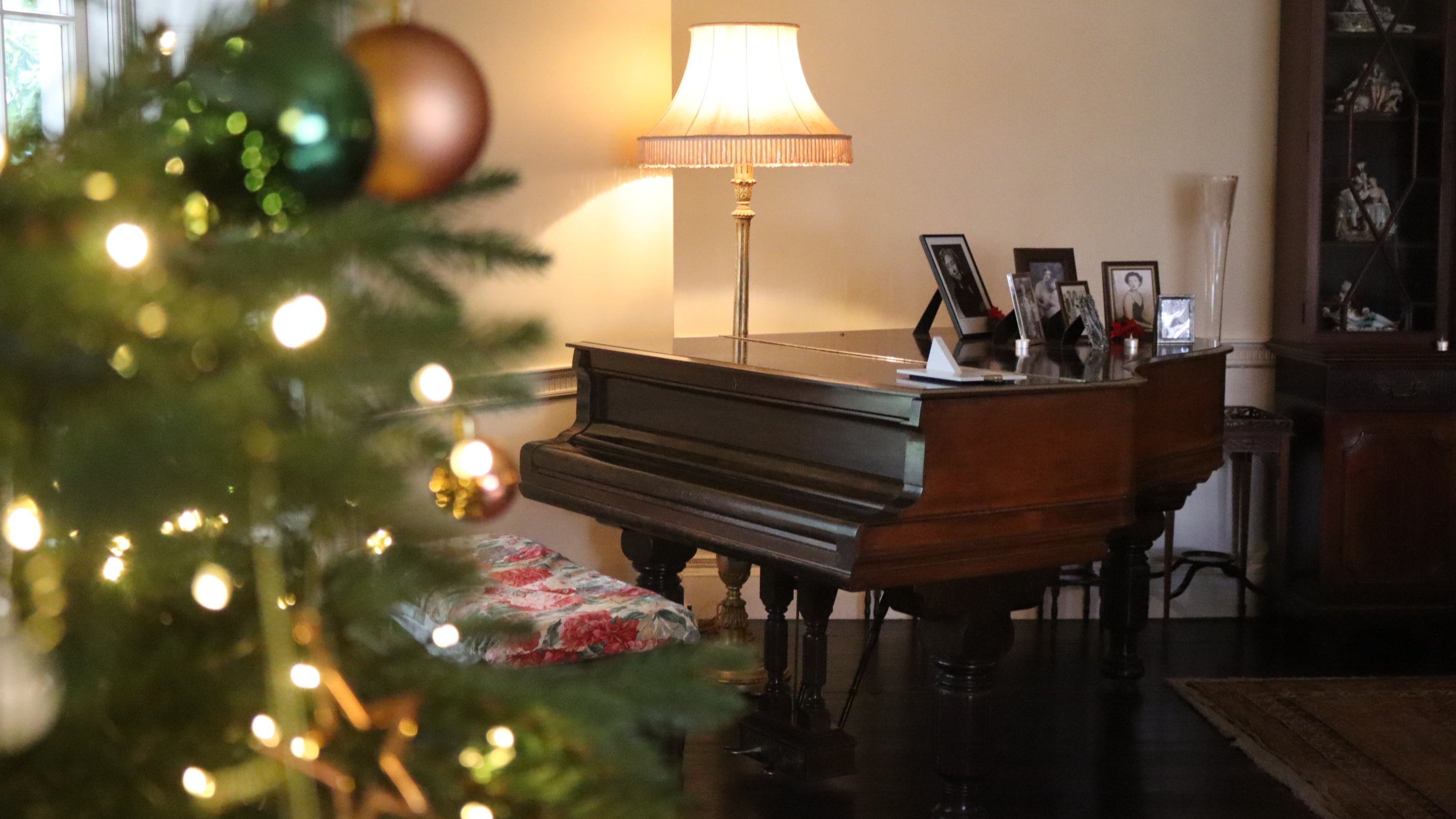 A wooden piano and stool in a room, with a lamp behind, framed by out of focus Christmas tree branches and fairy lights