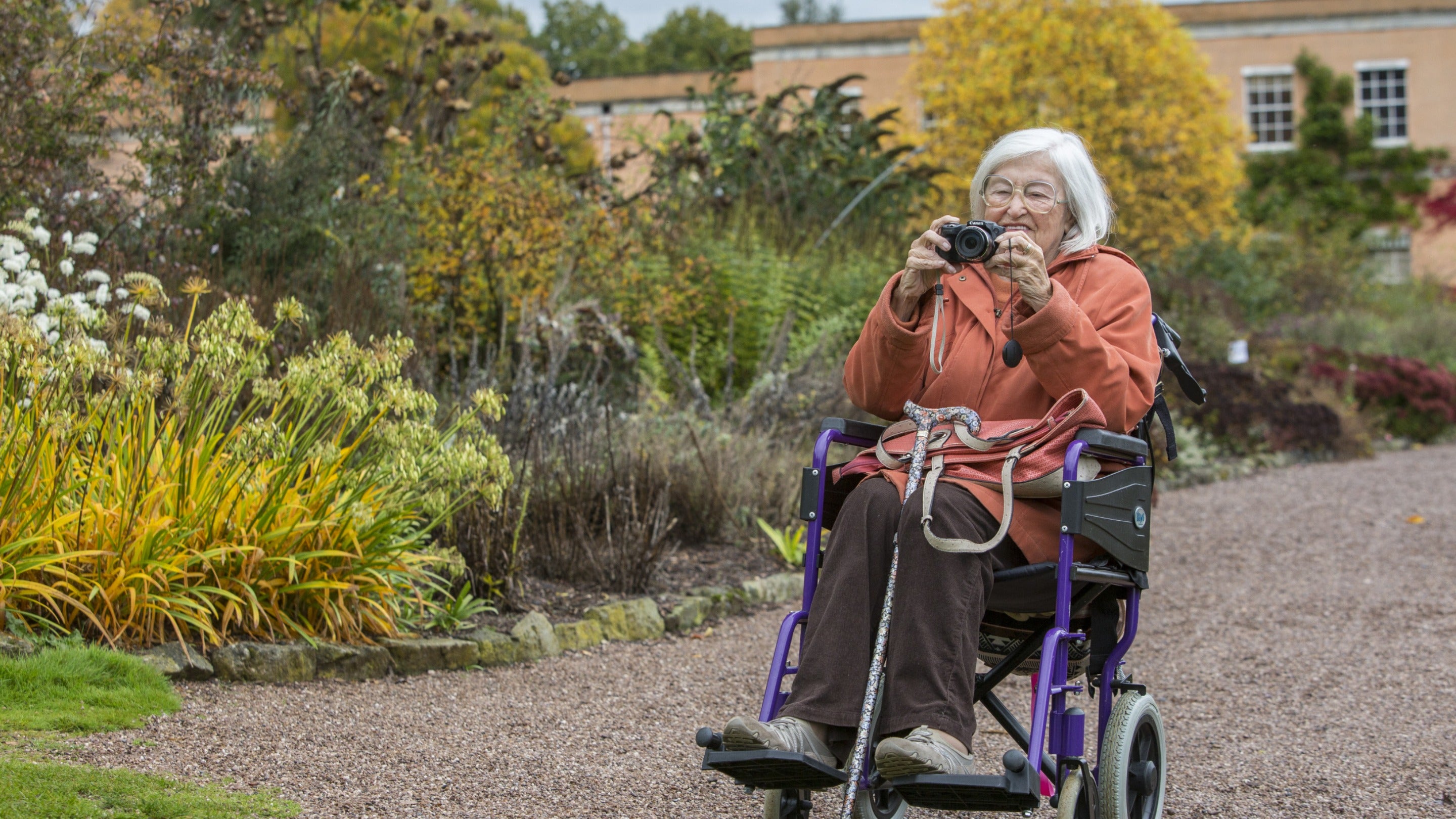 A visitor using a wheelchair takes a photo, with flower borders and Killerton house in the background