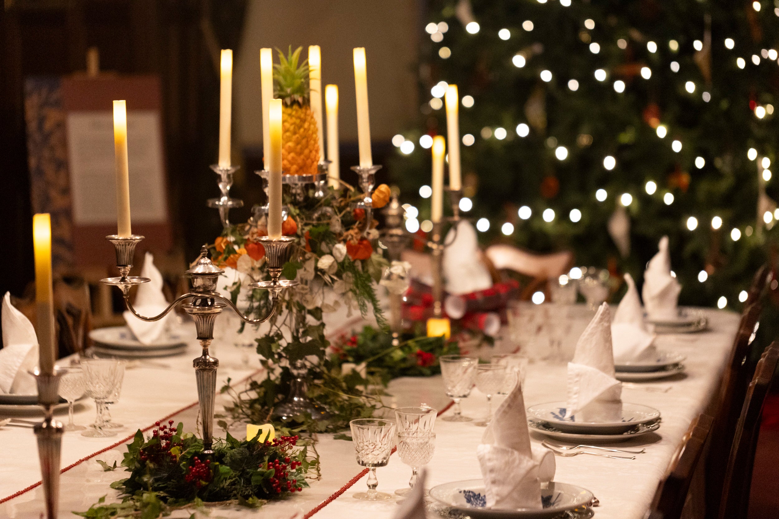 Festive dining table with cone-shaped napkins, crystal glasses, and a candelabrum topped with a pineapple, set against a lit Christmas tree.