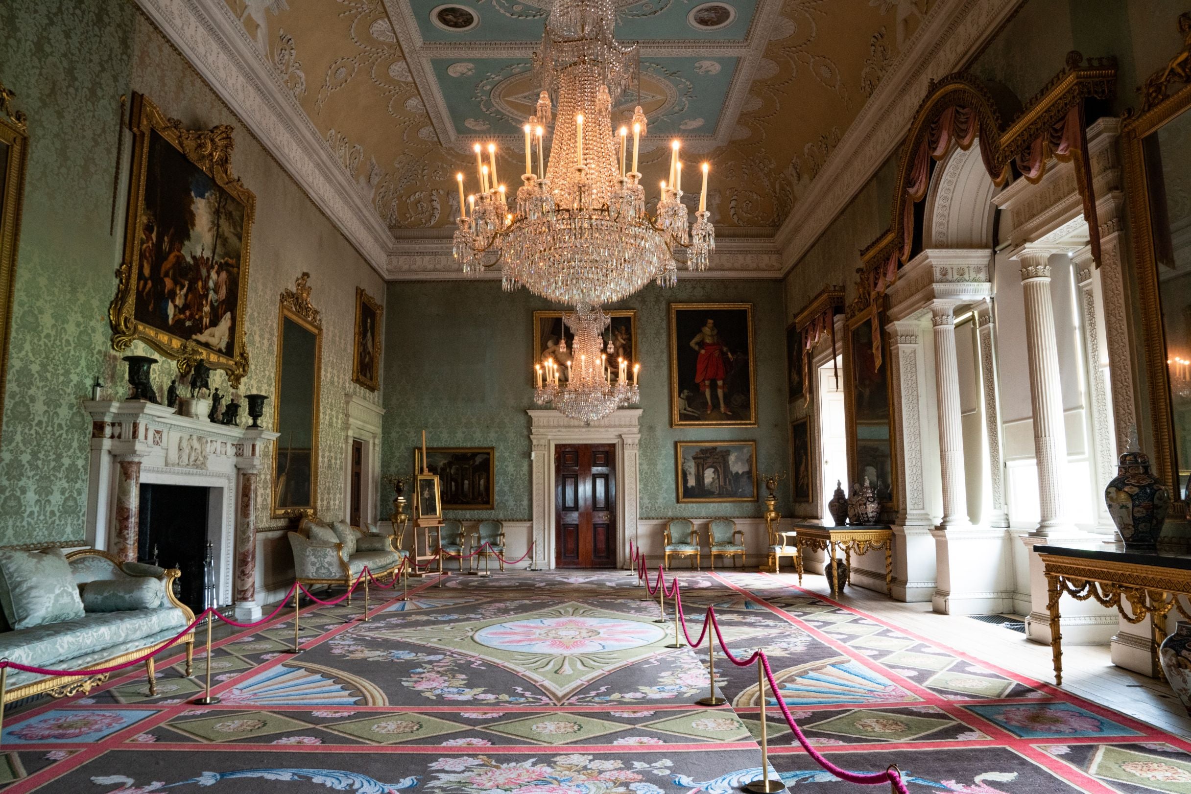 Saltram's luxurious Saloon with two lit chandeliers, damask wallpaper and a re-weave carpet covering half of the room so visitors can now walk through the space.