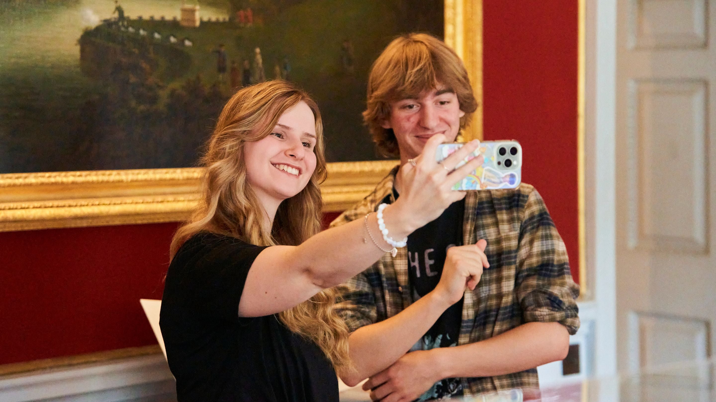 A teenage boy and girl take a selfie in front of a famous Tomkins painting inside an ornate red room at Saltram