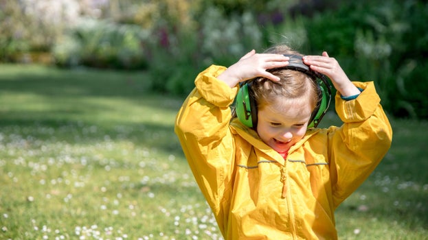 A girl wearing a yellow rain coat and wearing noise blocking headphones holds her ears and smiles