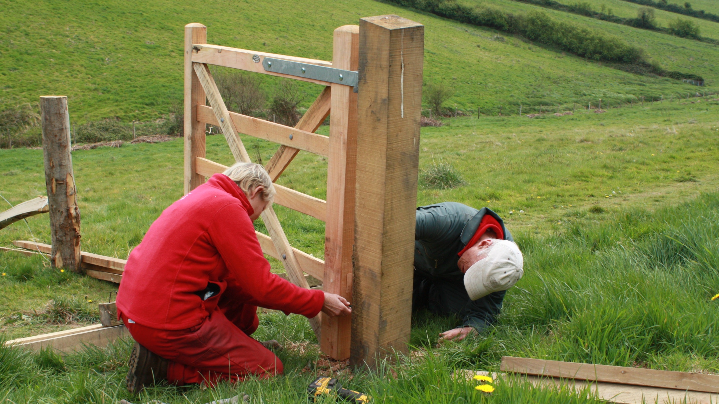 National Trust volunteers wearing red tops installing wooden gate posts in a green field.