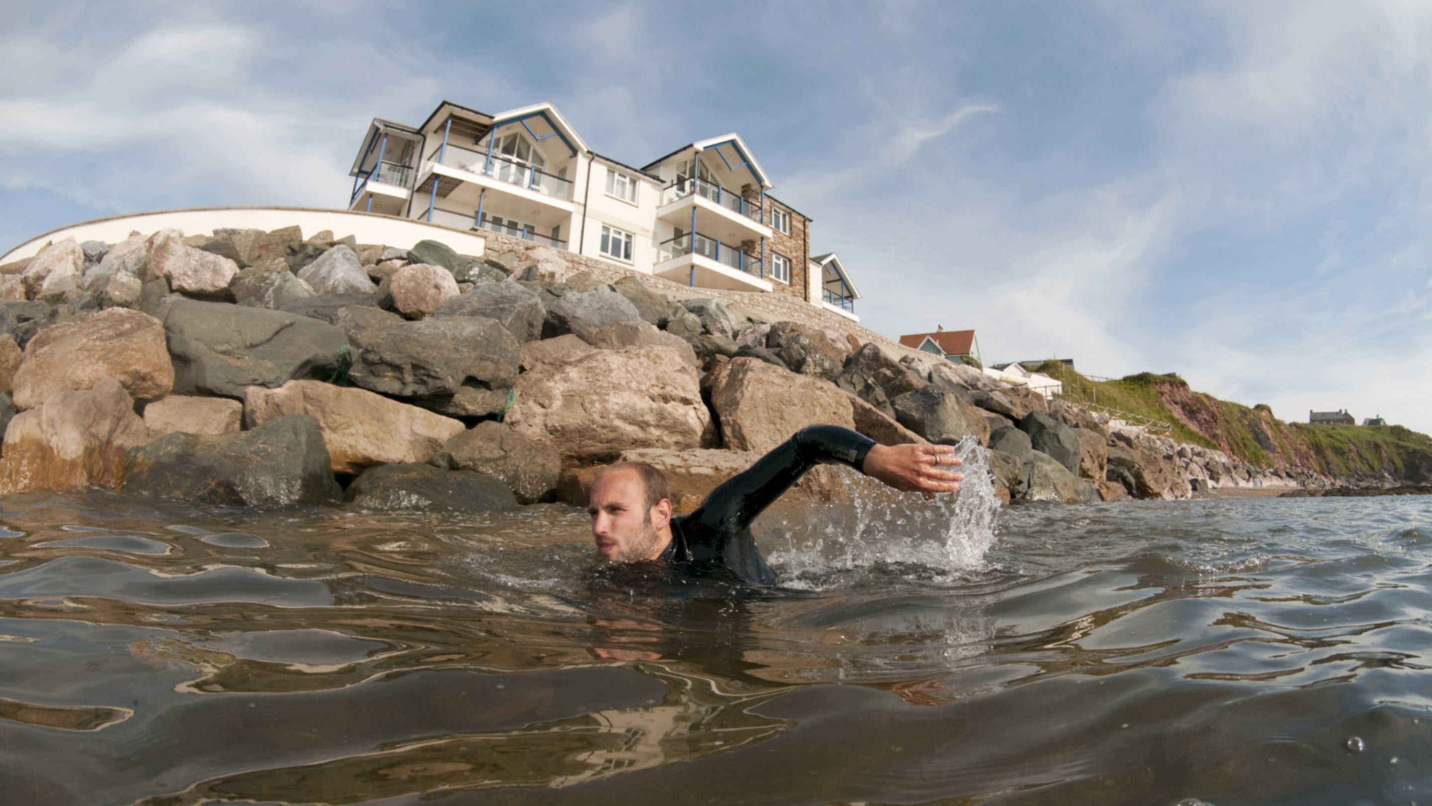Man in black wet suit swimming in the sea with rocks and house buildings in background