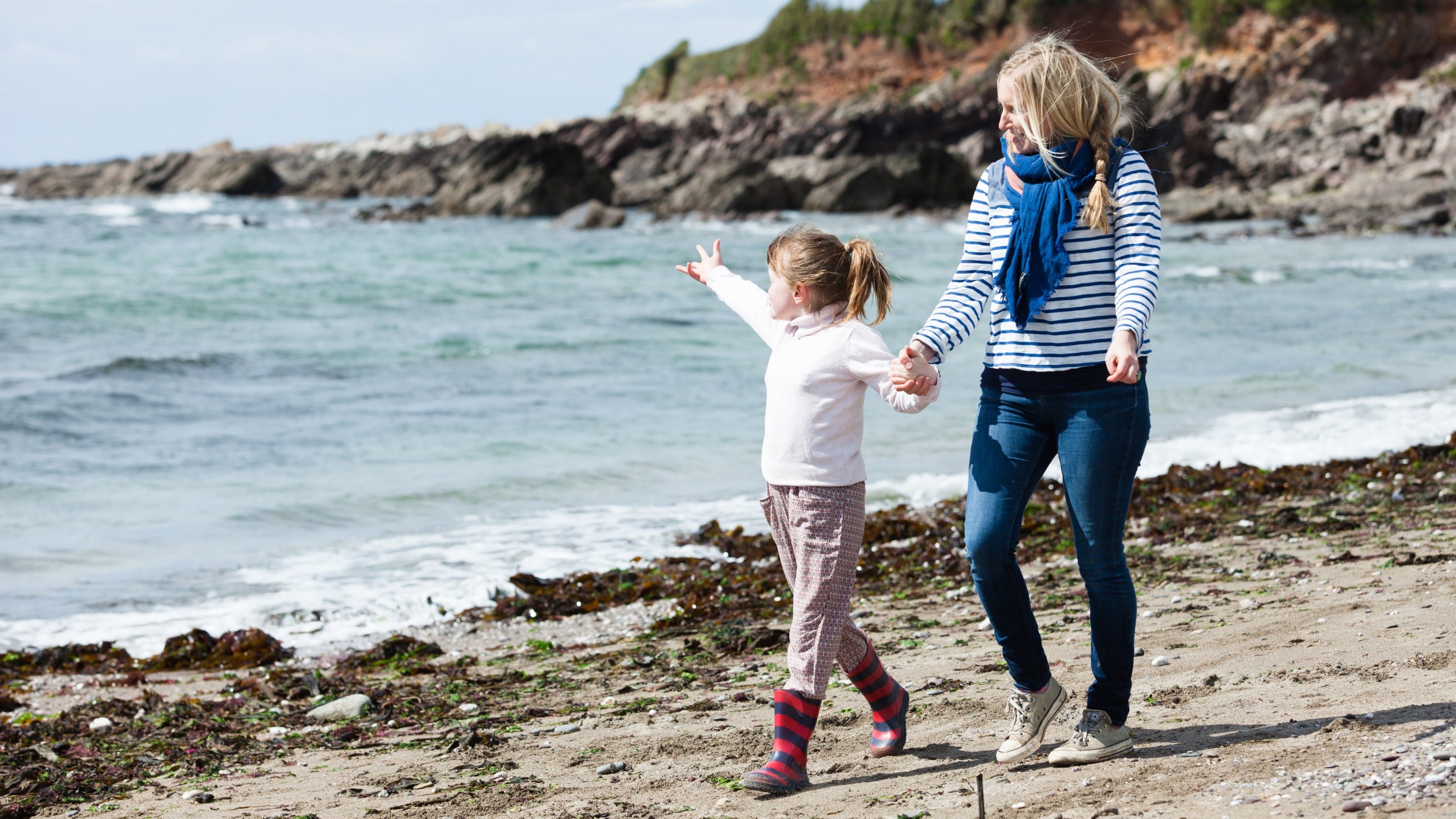 A woman and young girl walk along the shoreline in warm clothes, rocks in the background