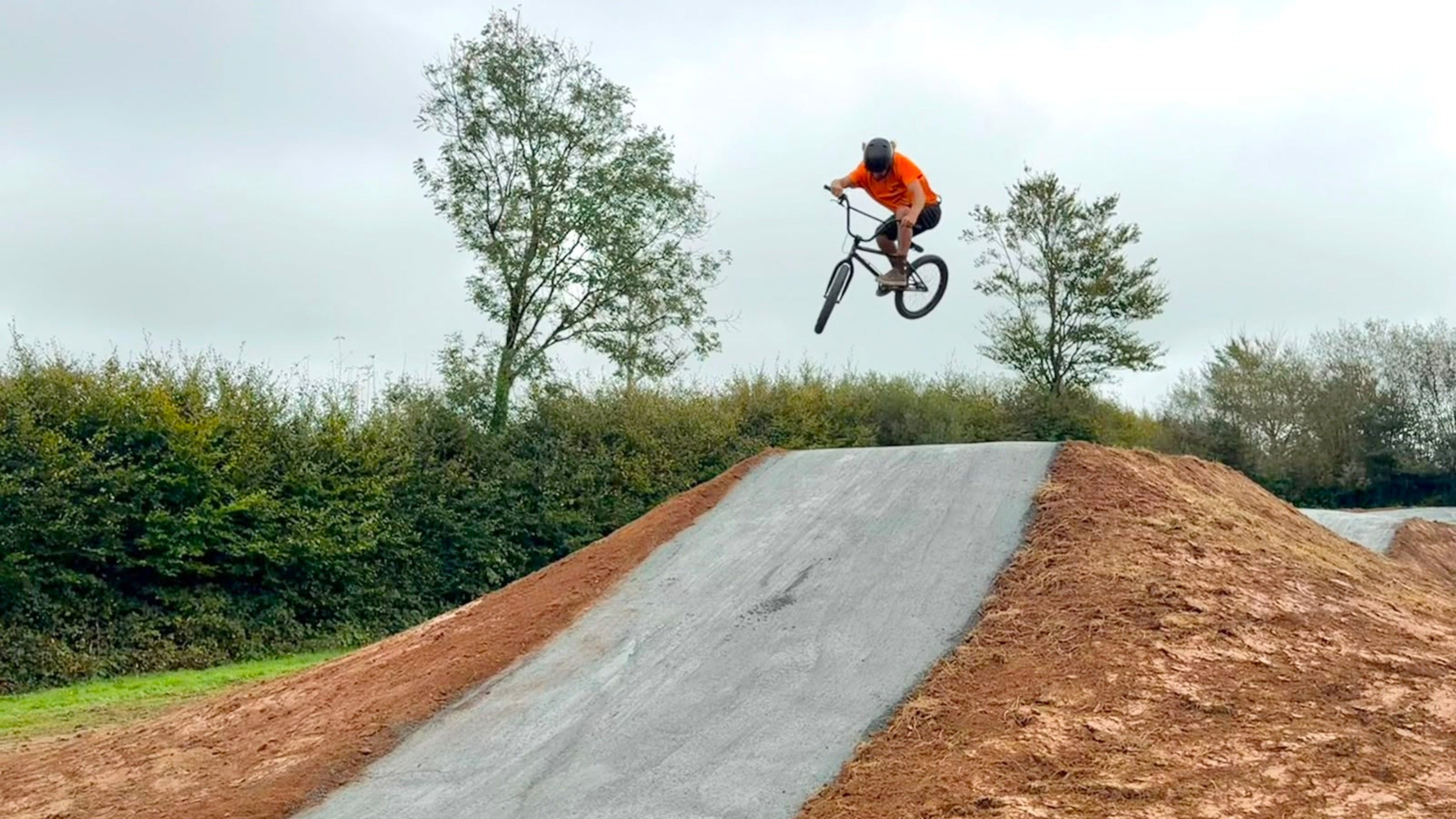 A bmx biker in the air over a box jump with a hedge and trees in the background