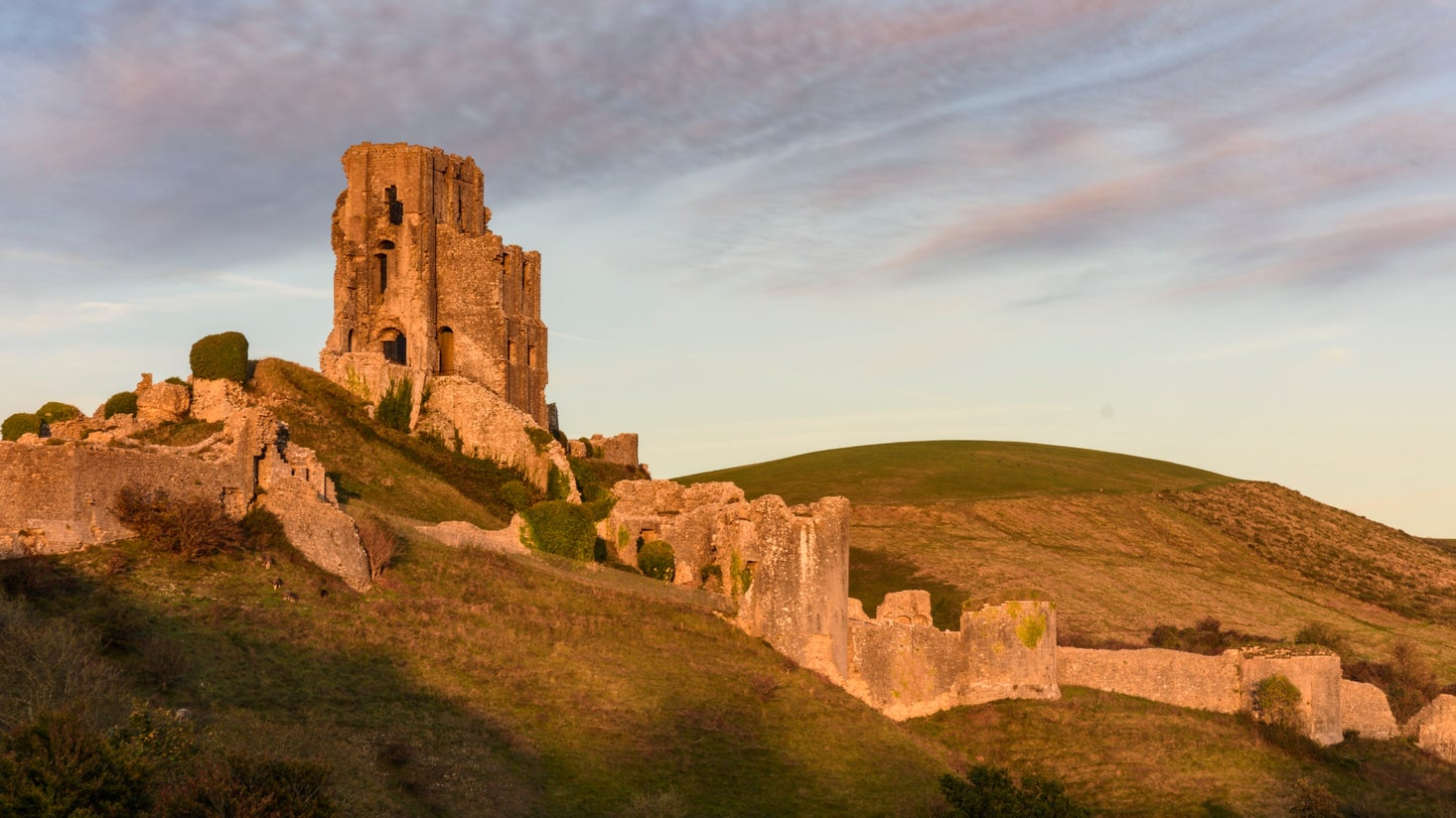 View of the ruins of Corfe Castle, lit in golden autumn sunlight, with a hill in the background